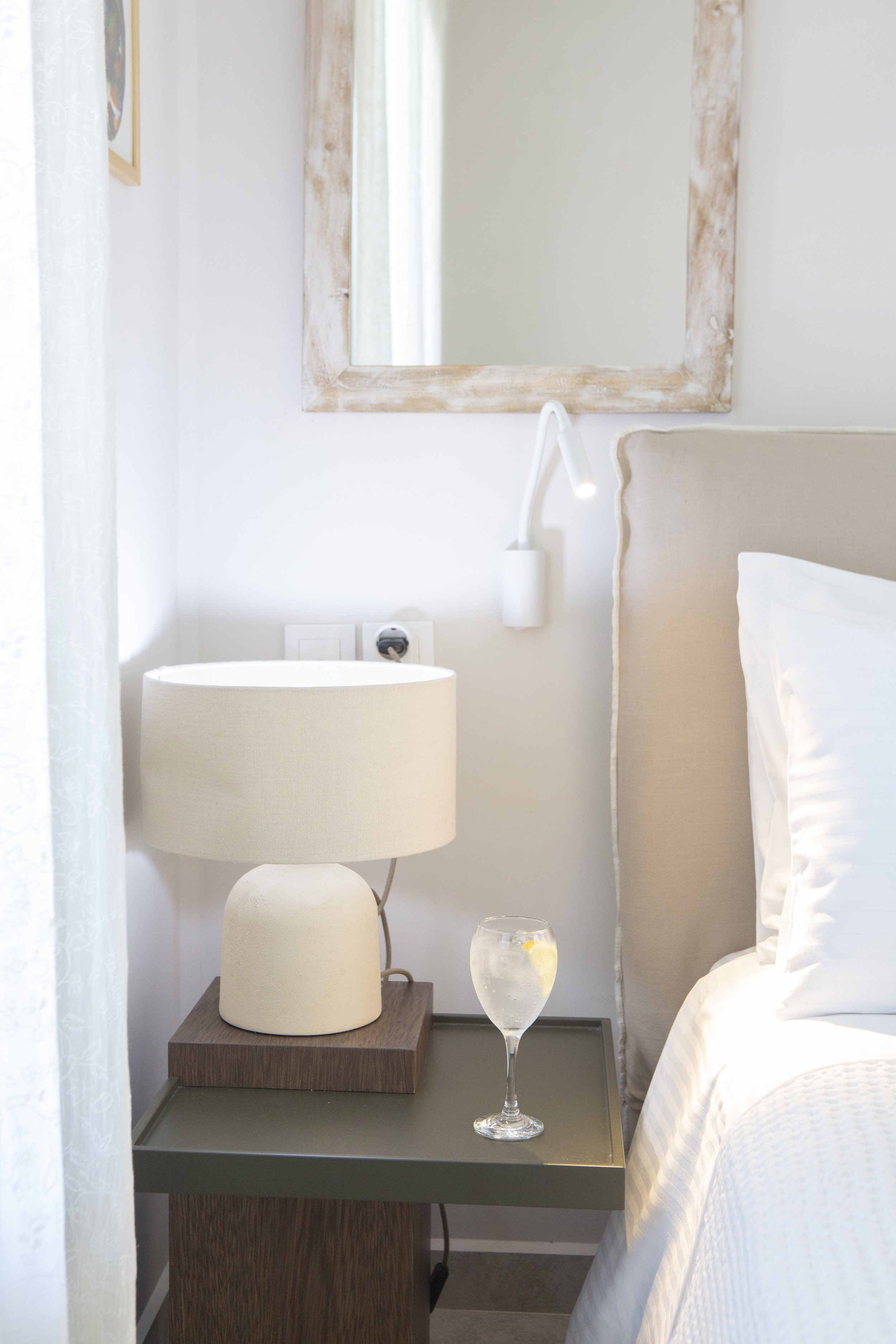 A hotel bedside table with a beige lampshade on a white lamp, a glass of water with lemon slices, and a wooden base, next to a bed with white linens and a beige headboard, and a white wall with a mirror and a wall-mounted lamp.