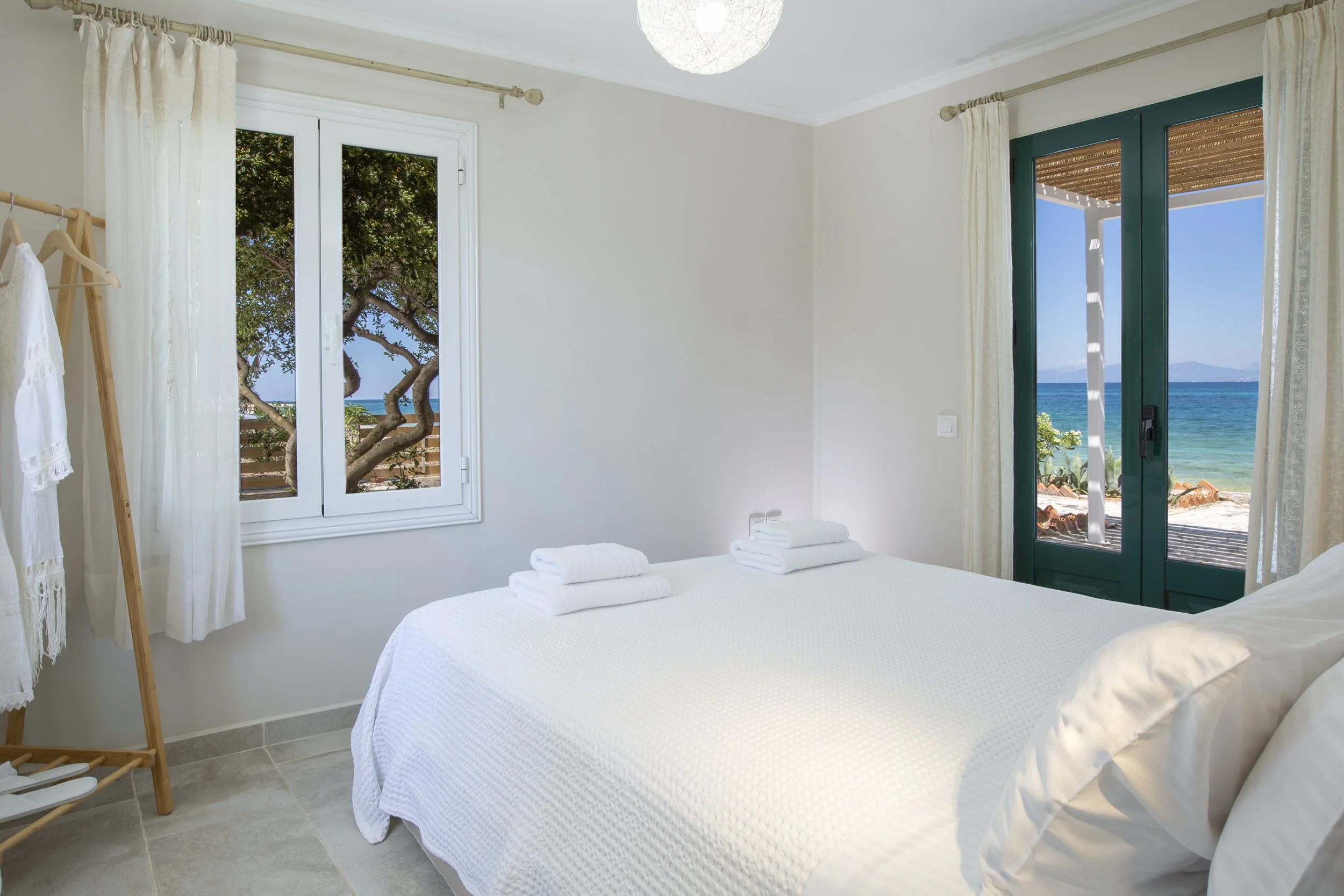 A bedroom with a large bed covered in a white textured bedspread and folded towels on top. There are two windows, one showing a view of trees and the beach, and another with a door leading outside to a balcony with a bamboo shade. The room has light-