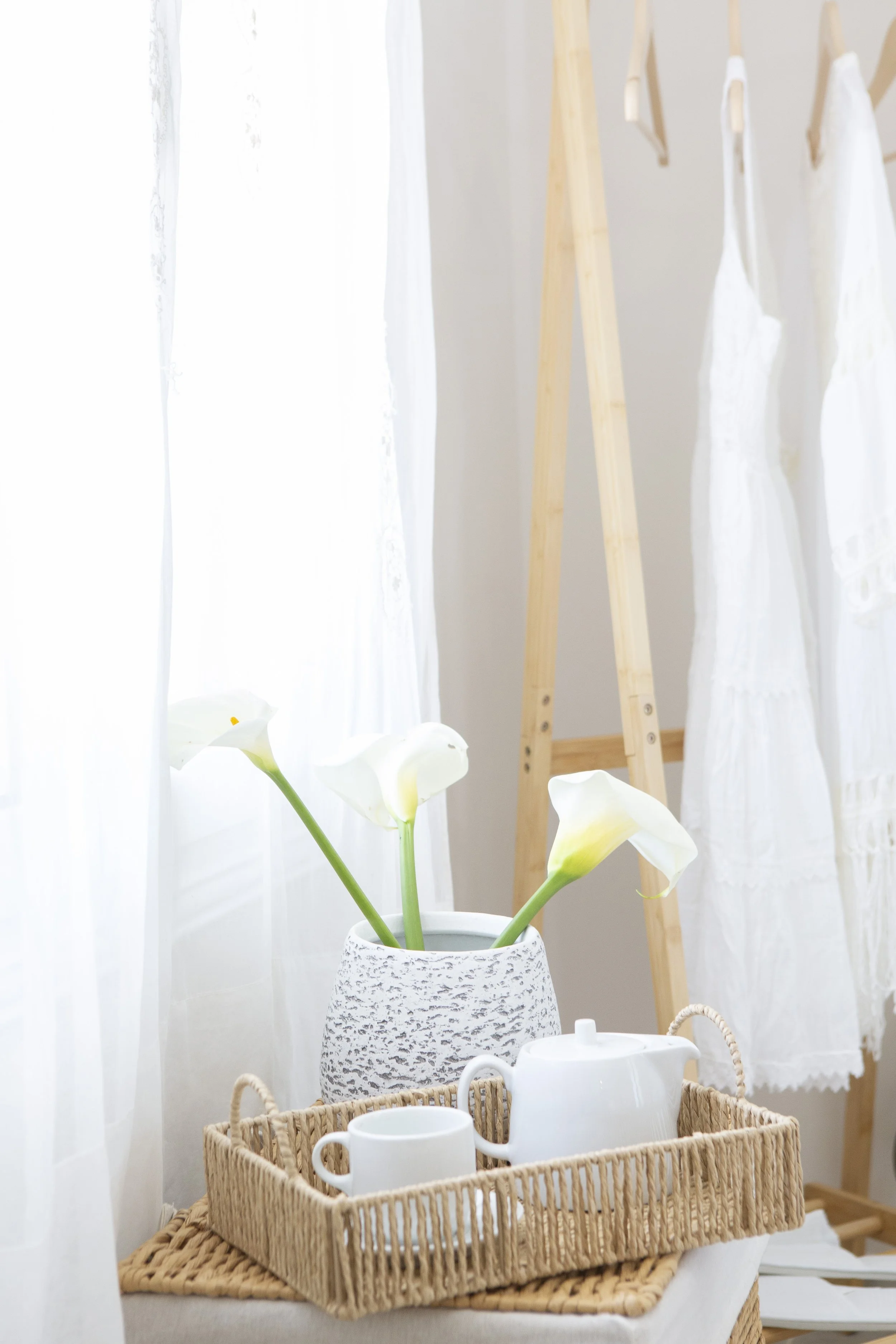 A cozy indoor scene featuring a white plant in a textured vase, a wooden laundry drying rack with white clothes hanging, and a wicker tray with white teapot, cups, and a jug, all near white curtains allowing soft light to fill the space.
