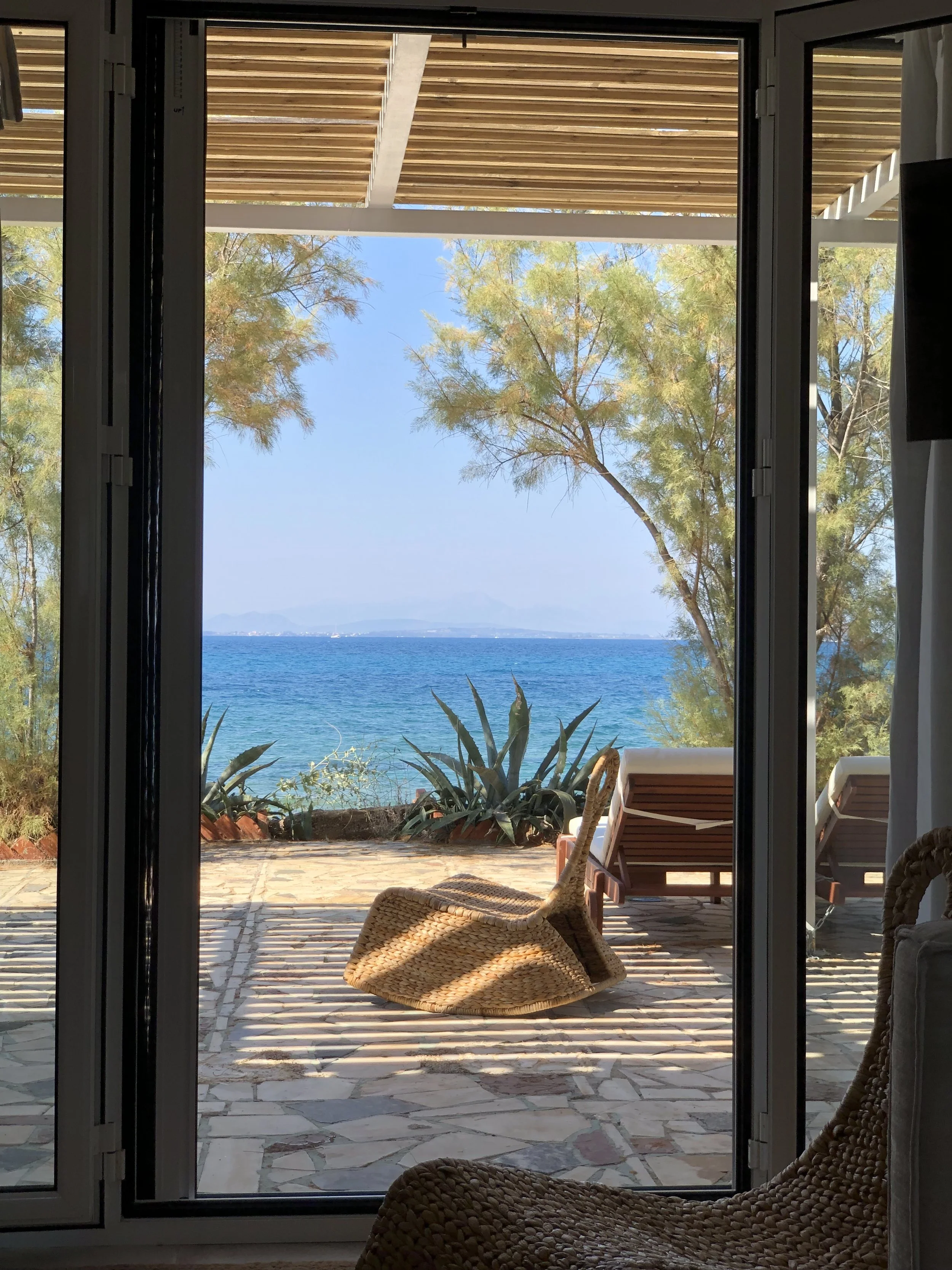 Photo of an ocean view through open glass doors, with a stone patio, wicker chairs, two lounge chairs, and green plants, under a wooden roof.
