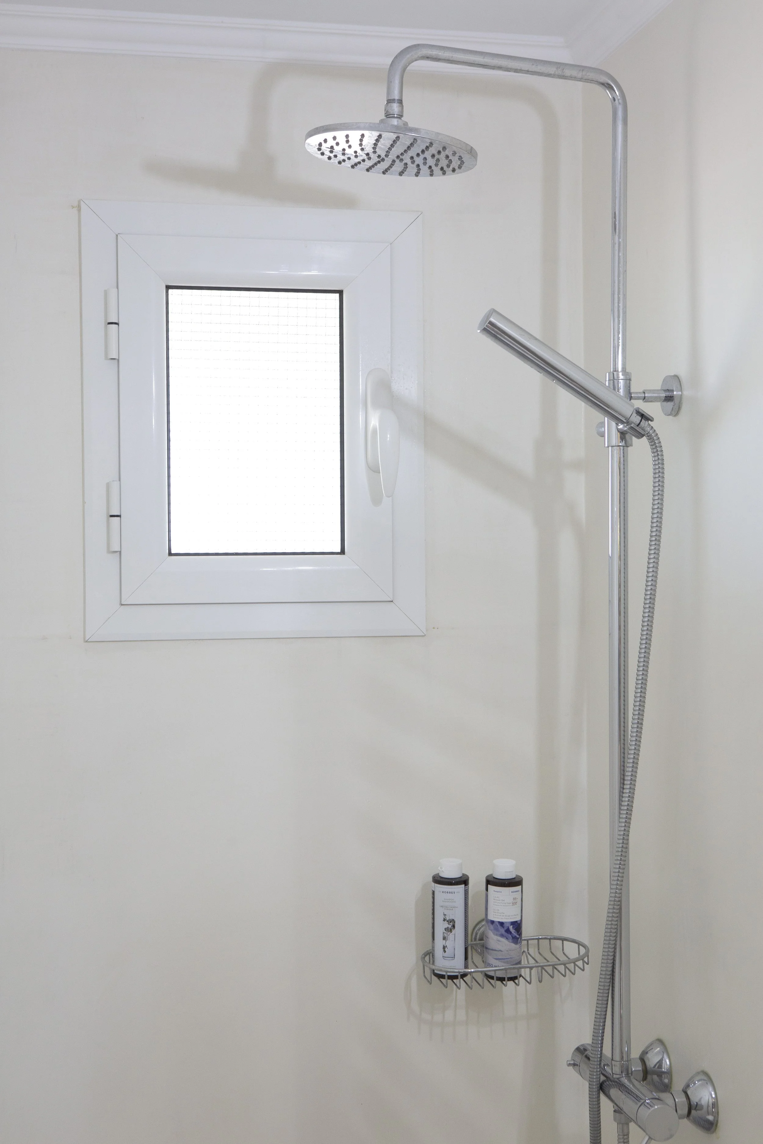 Shower with a round showerhead, a handheld shower attachment, a window with frosted glass, and two bottles of toiletries on a wire shelf.