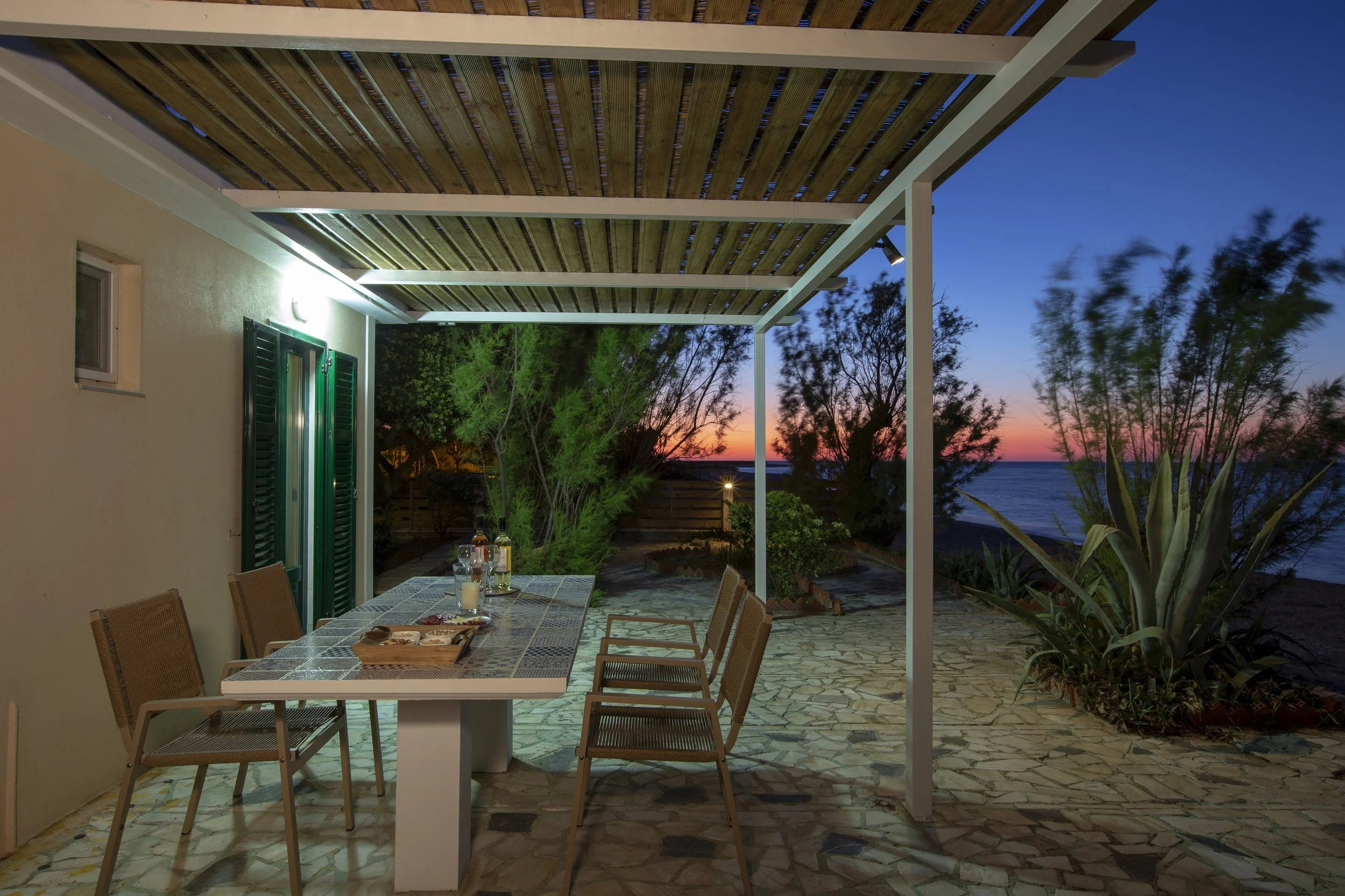 Outdoor patio area with dining table and chairs, decorated with drinks and snacks, set under a pergola at dusk with view of trees and ocean in the background.