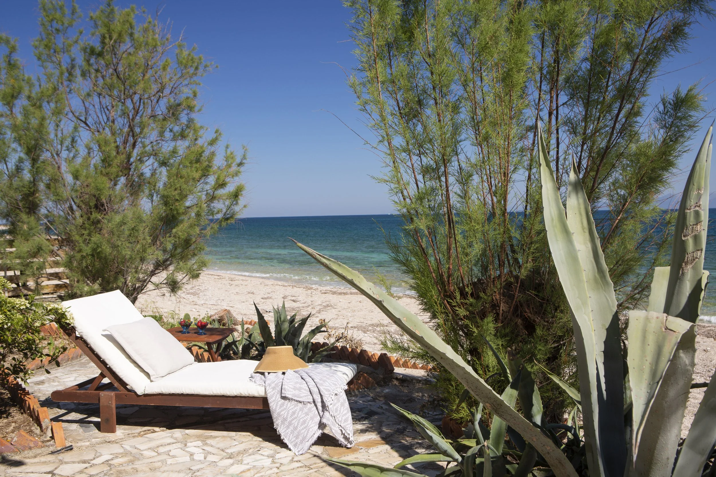 Beach scene with white lounge chairs, small table with colorful glasses, and large green plants including agave and cypress trees, overlooking the ocean with a clear blue sky.