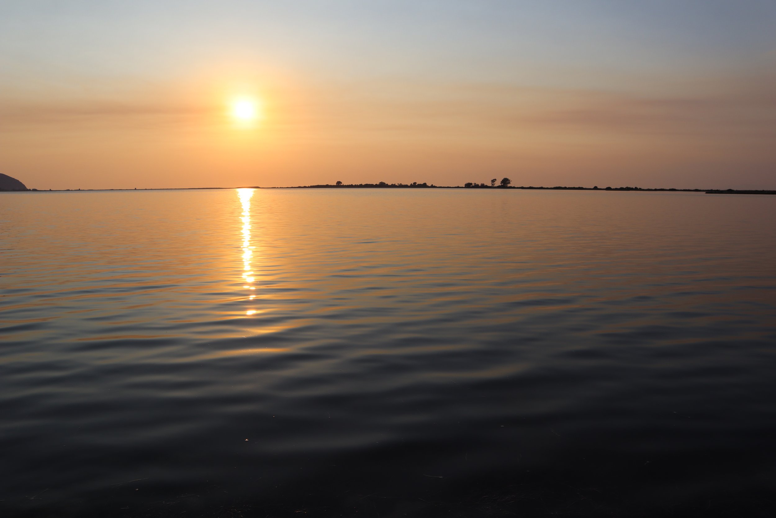 Sunset over calm water with a reflection of the sun on the surface, a distant shoreline with trees in the background.