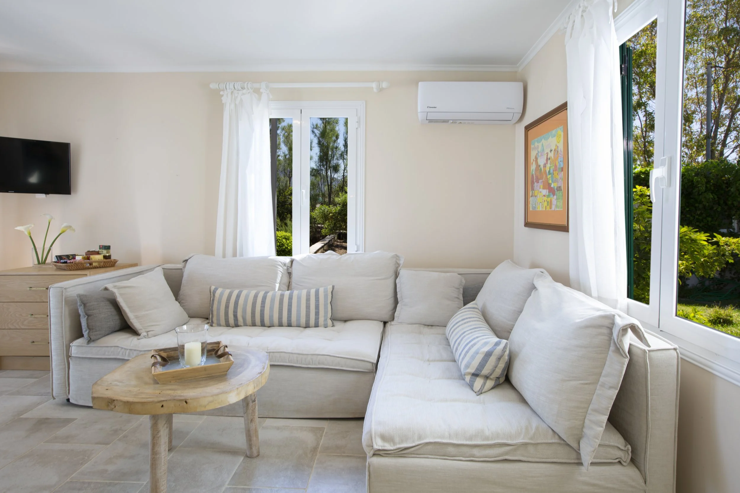 Living room with white sofa, striped pillows, coffee table with candles, TV on wall, windows with white curtains, and view of trees outside.
