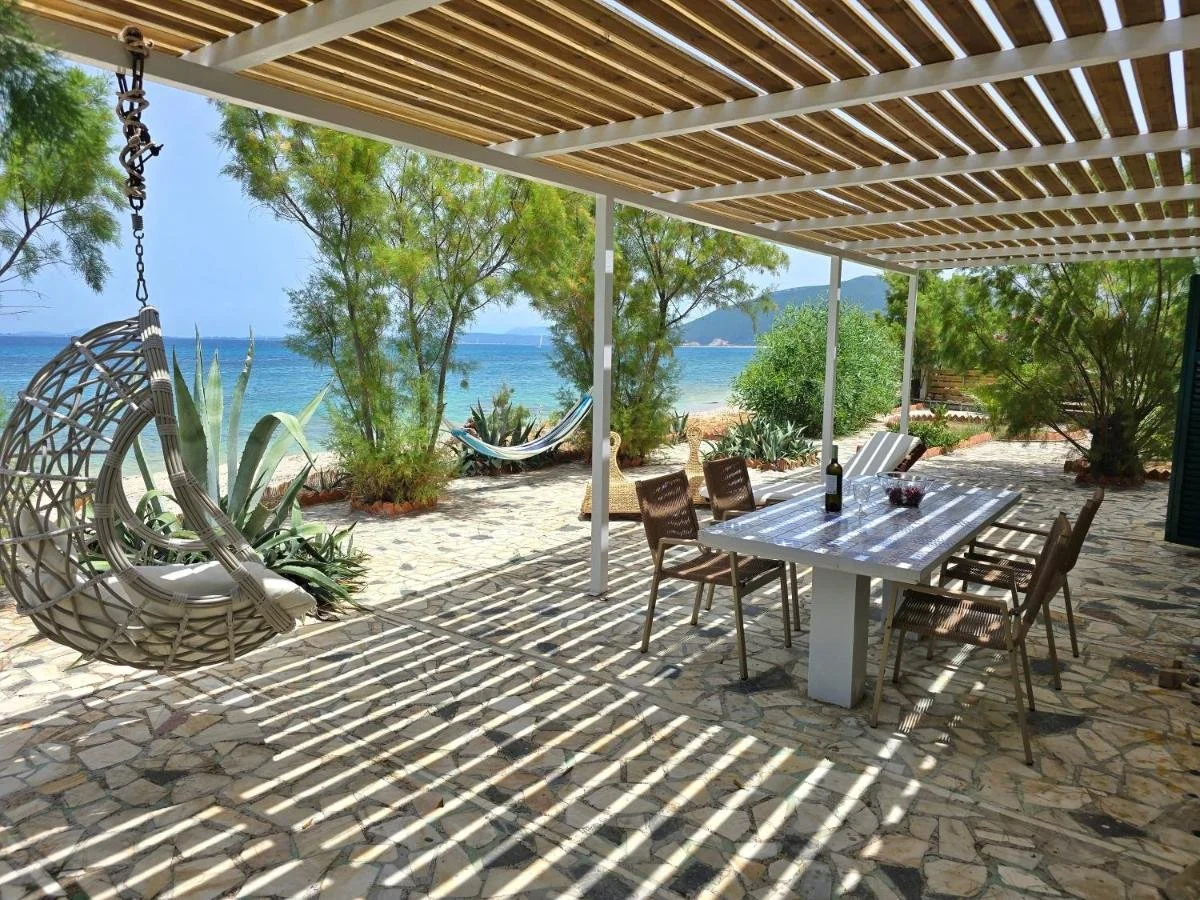 Beachside patio with a hanging egg chair, a white dining table with chairs, a bottle of wine, and a view of the ocean, trees, and a hammock in the background.
