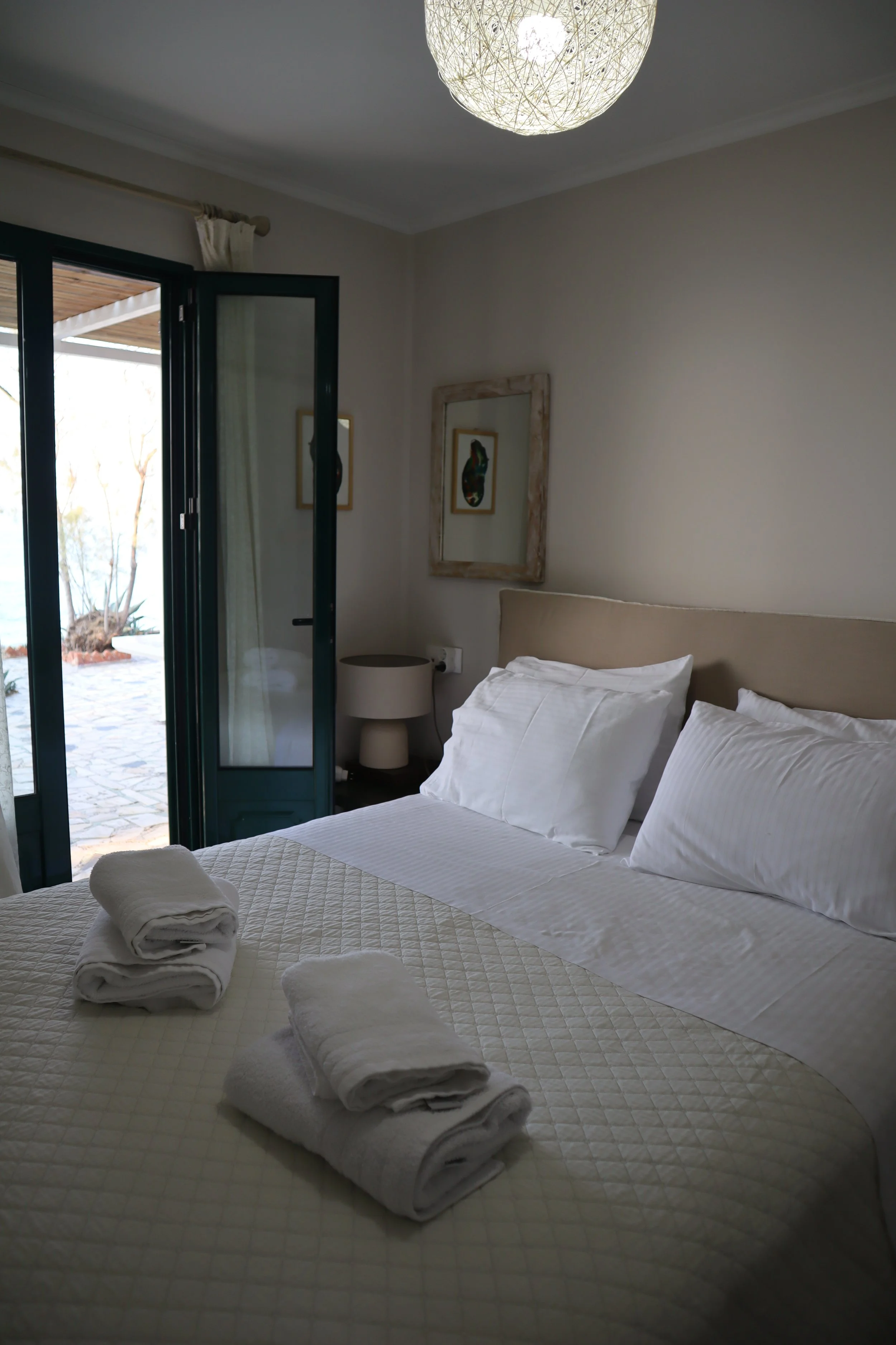 A neatly made bed with white linens and folded towels on top, in a bright bedroom with a large window and door open to a patio, with artwork on the walls and a round ceiling light.