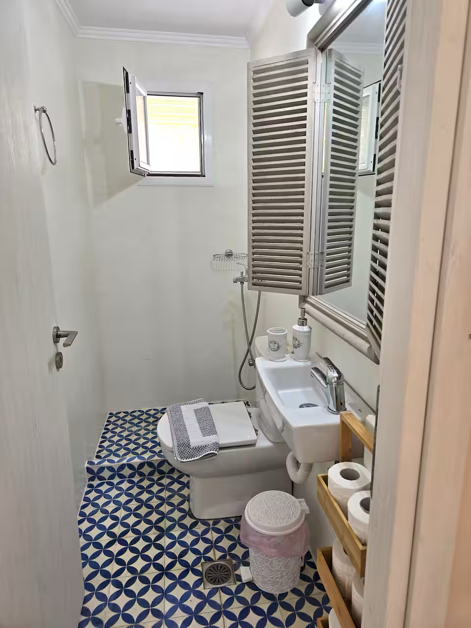 Small bathroom with a toilet, a small sink, wooden shelves with toilet paper rolls, a trash can with a pink liner, a mirror, and a small window with shutters.
