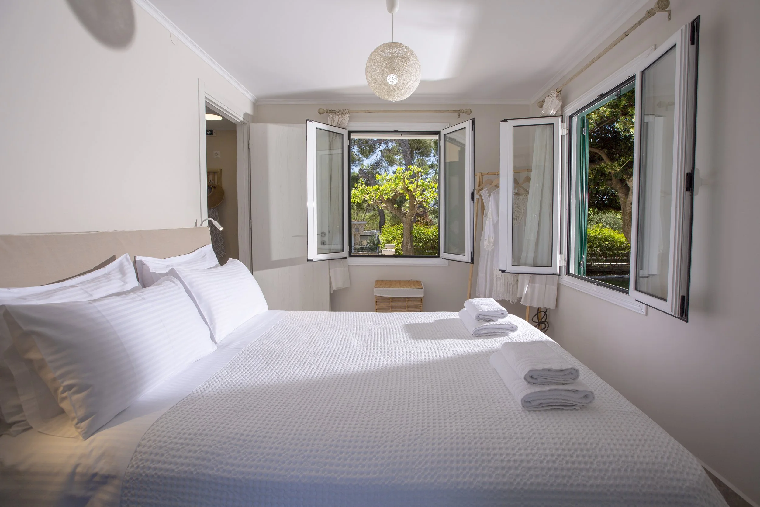 A bright, cozy bedroom with white walls and bedding, open windows showing green trees outside, and neatly folded towels on the bed.