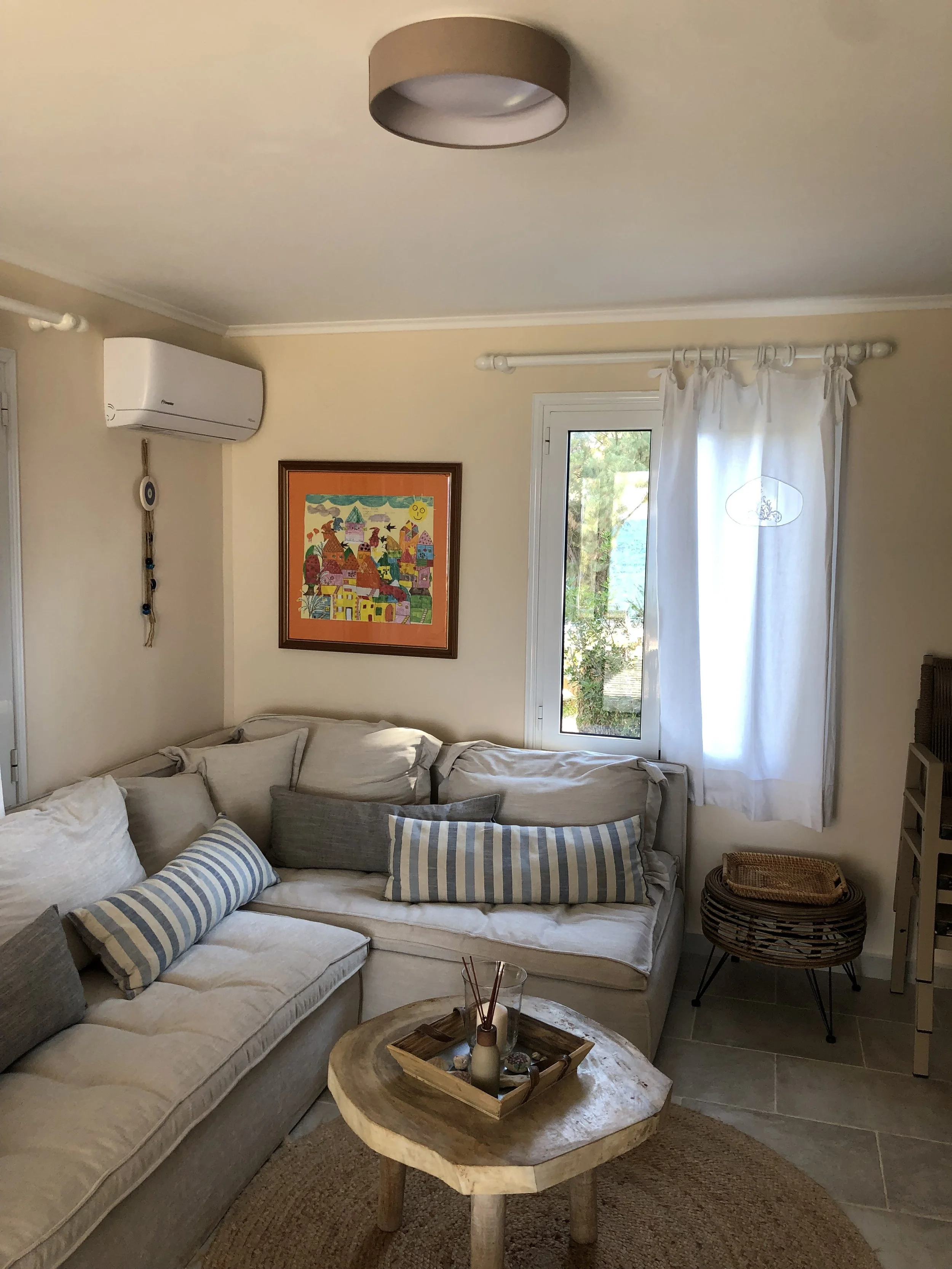 Living room with beige sofa, colorful artwork on the wall, a window with white curtains, and a wooden coffee table with decorative items.