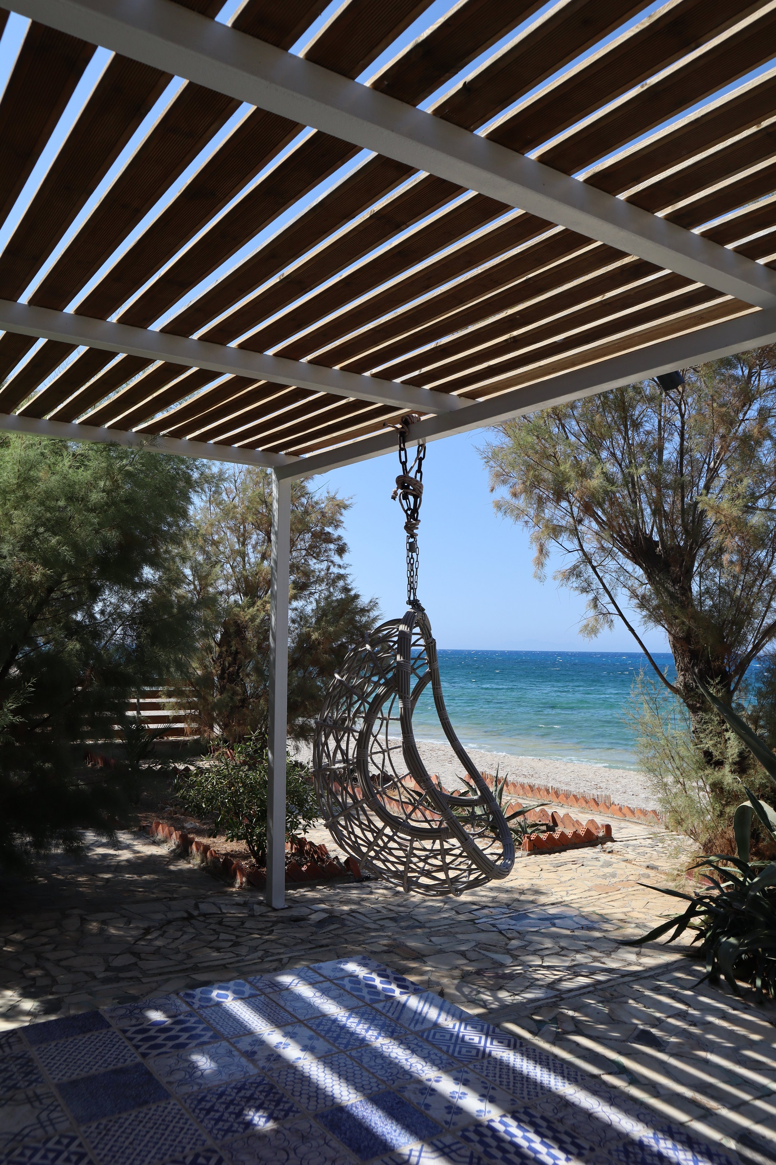 A hanging wicker chair on a porch overlooking the beach and ocean, with trees and a stone pathway, under a wooden pergola.