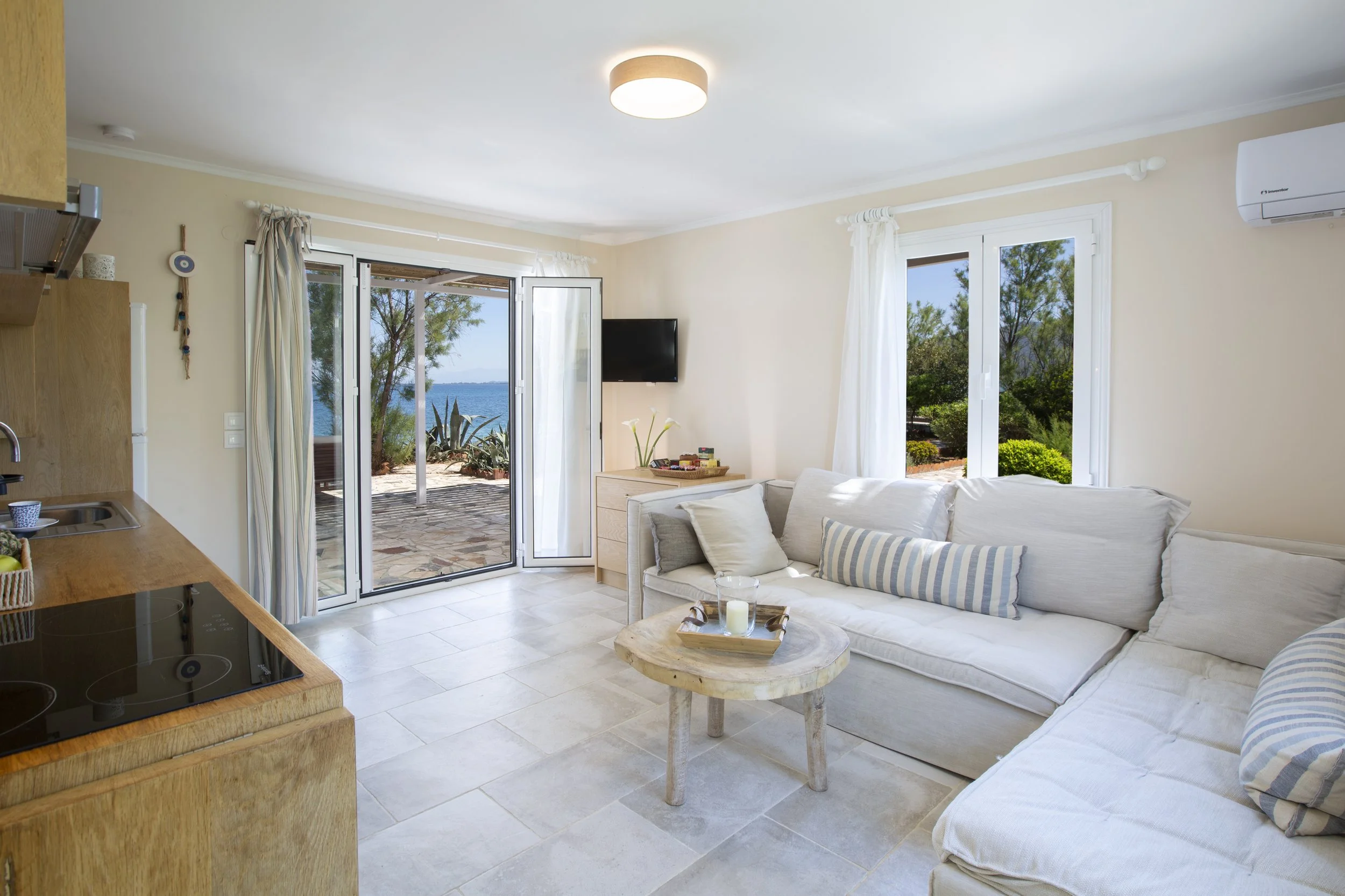 Living room with beige walls and large windows showing a view of trees and water, white curtains, a white sofa with striped pillows, a wooden coffee table with candles, a wall-mounted TV, and an open balcony door.