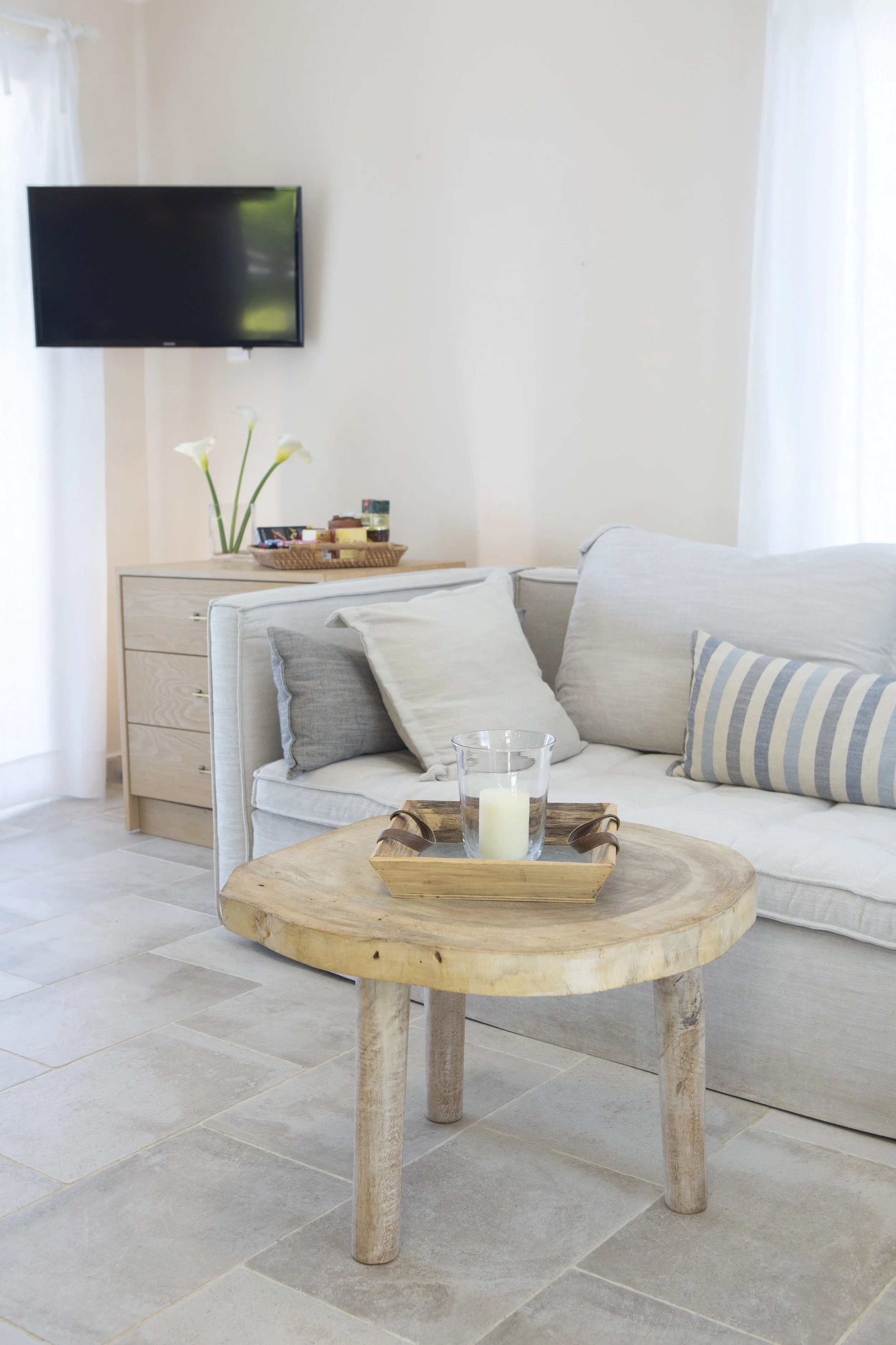 Living room with a wooden coffee table holding a glass candle in a glass holder on a tray, a sofa with cushions, a dresser with a basket, flowers, and snacks, and a wall-mounted TV, with natural light coming through curtains.
