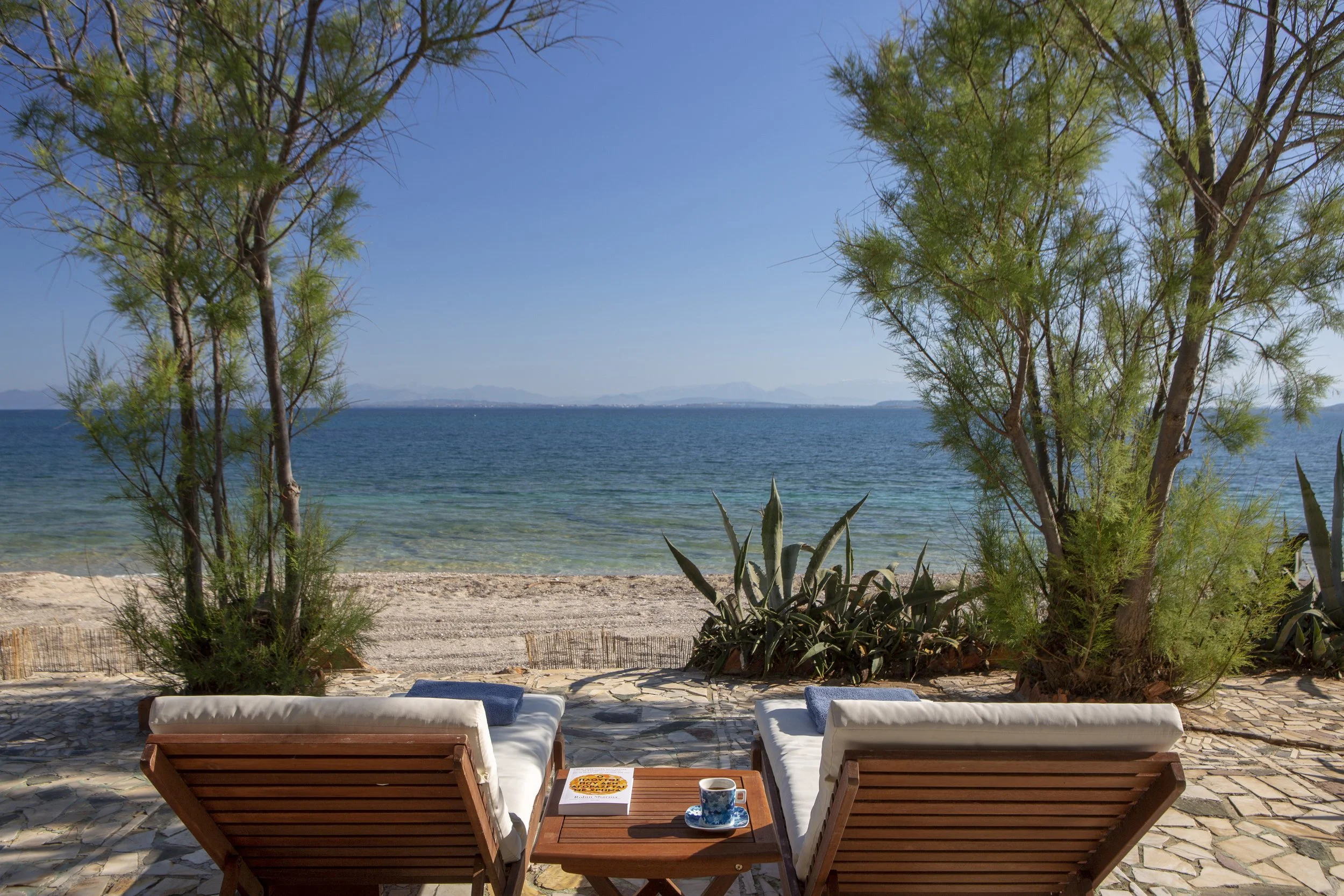 Beach scene with two lounge chairs facing the water, small table with a book and a coffee mug, framed by trees and shrubs, with a view of the ocean and distant mountains under a clear blue sky.