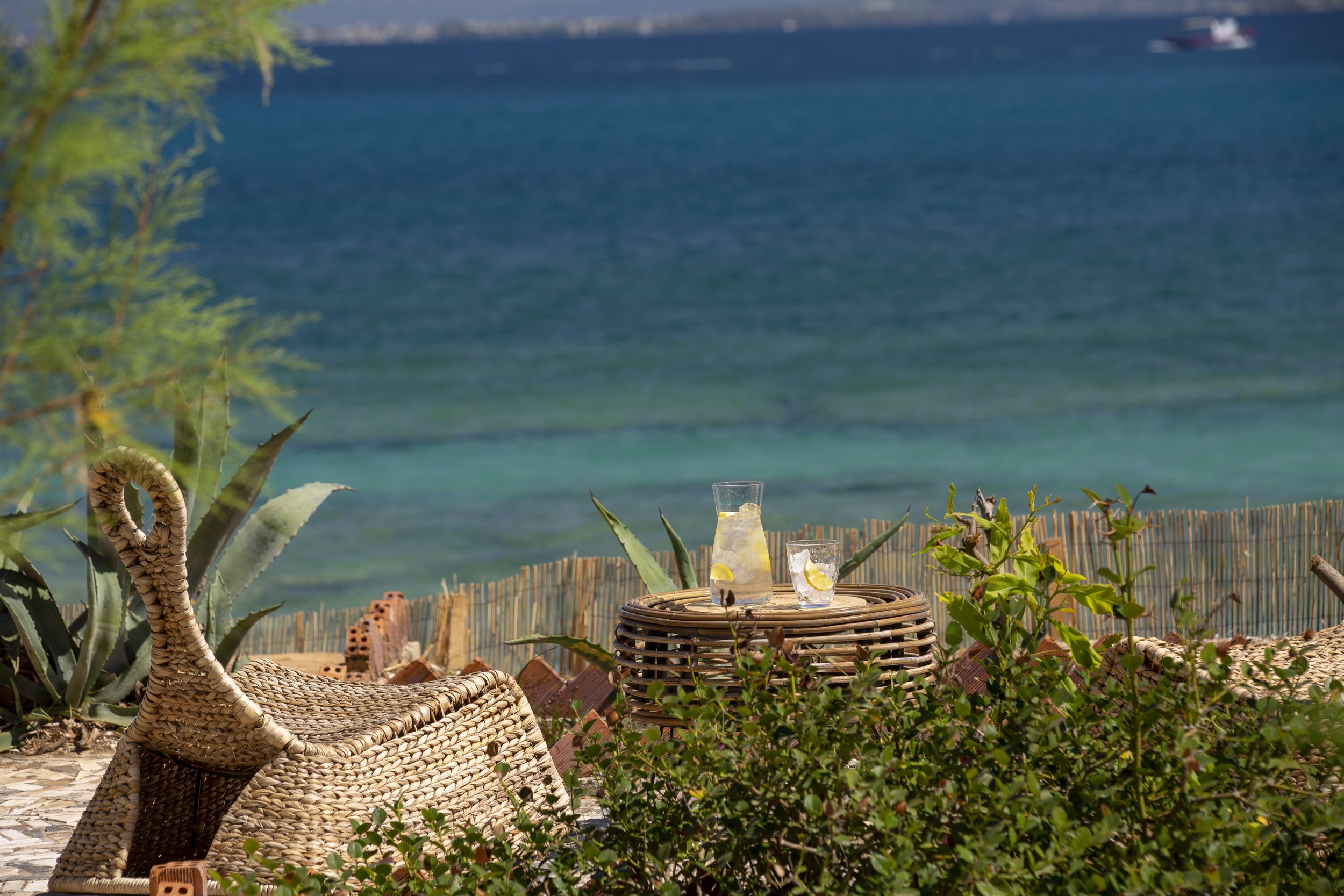 Outdoor patio scene with wicker chairs, a small round table with a pitcher and glass of lemon water, surrounded by plants, overlooking the ocean with a distant ship