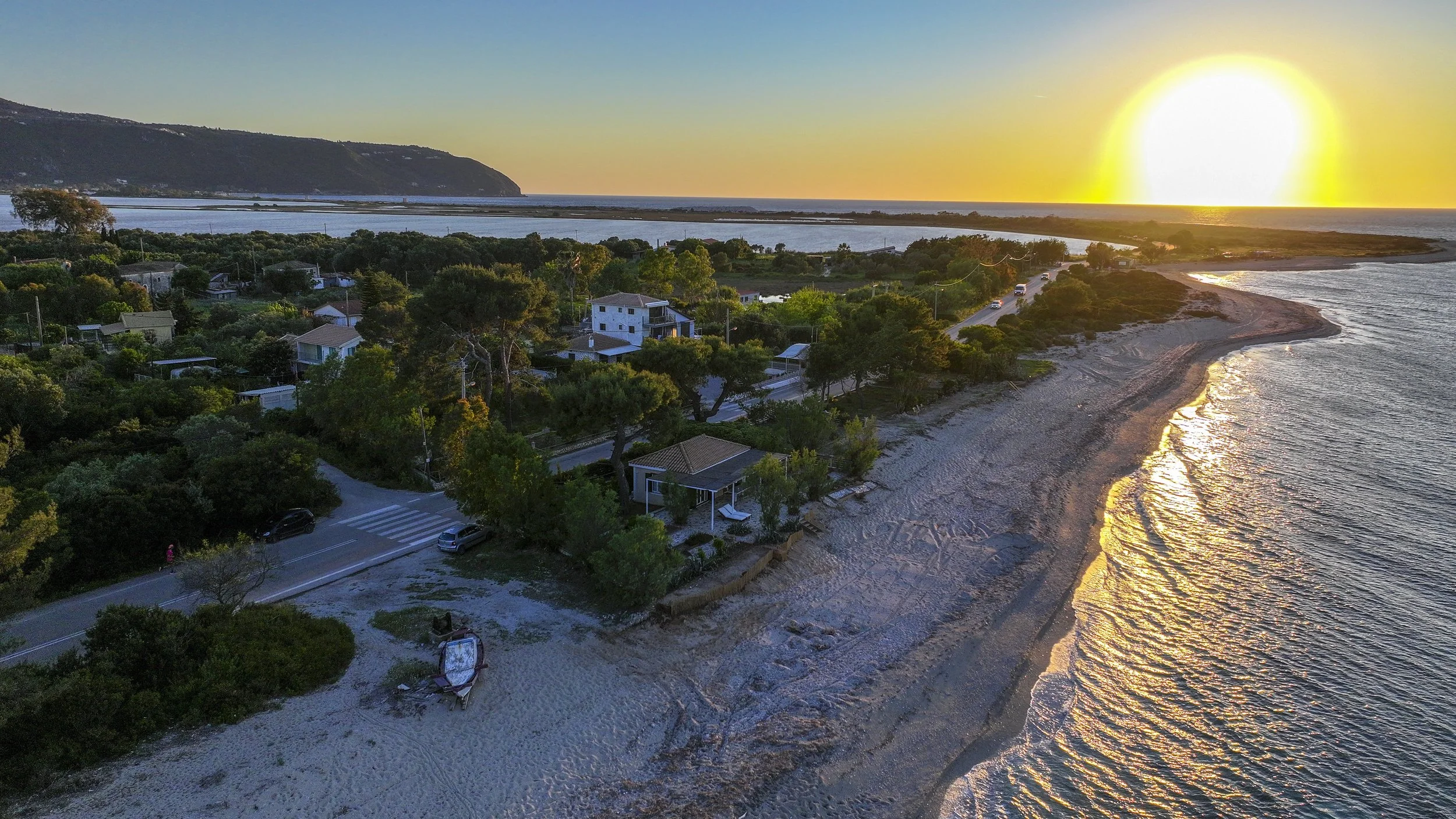 A coastal scene at sunset showing a sandy beach, a peaceful ocean, green trees, houses, and a mountain in the background.