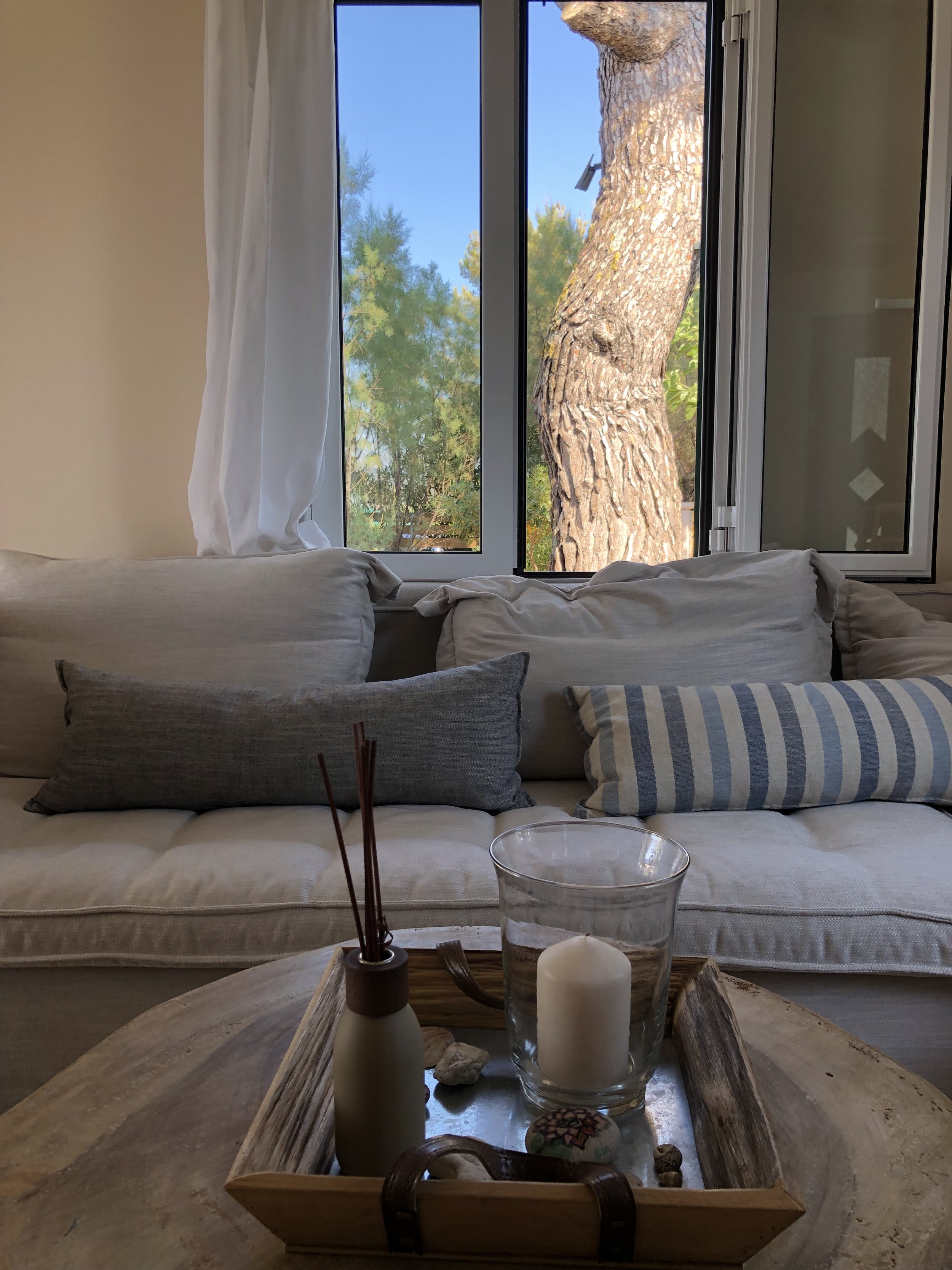 Living room with cream-colored sofa, decorative pillows, and a wooden coffee table with candles, rocks, and a reed diffuser, next to a window showing a tree outside.