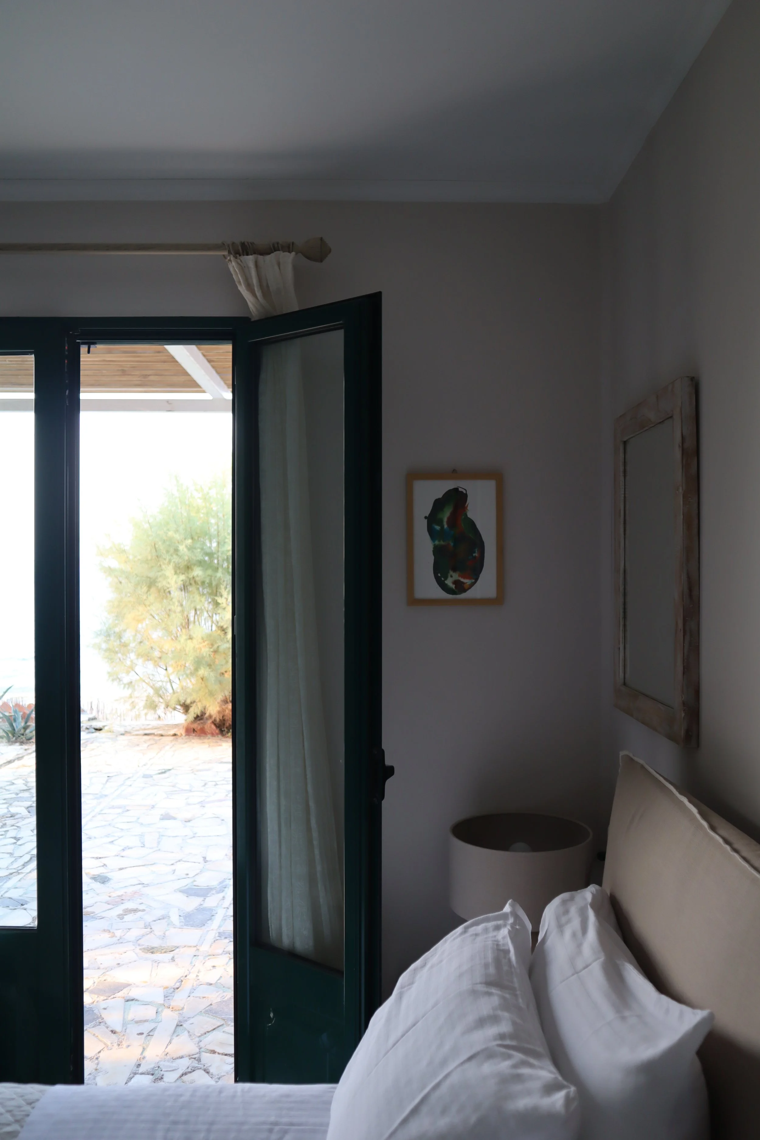 A bedroom with a partially opened door leading to an outdoor patio with stone flooring and a shrub. Inside, there is a bed with white bedding, a nightstand with a lamp, and artwork hanging on the wall.