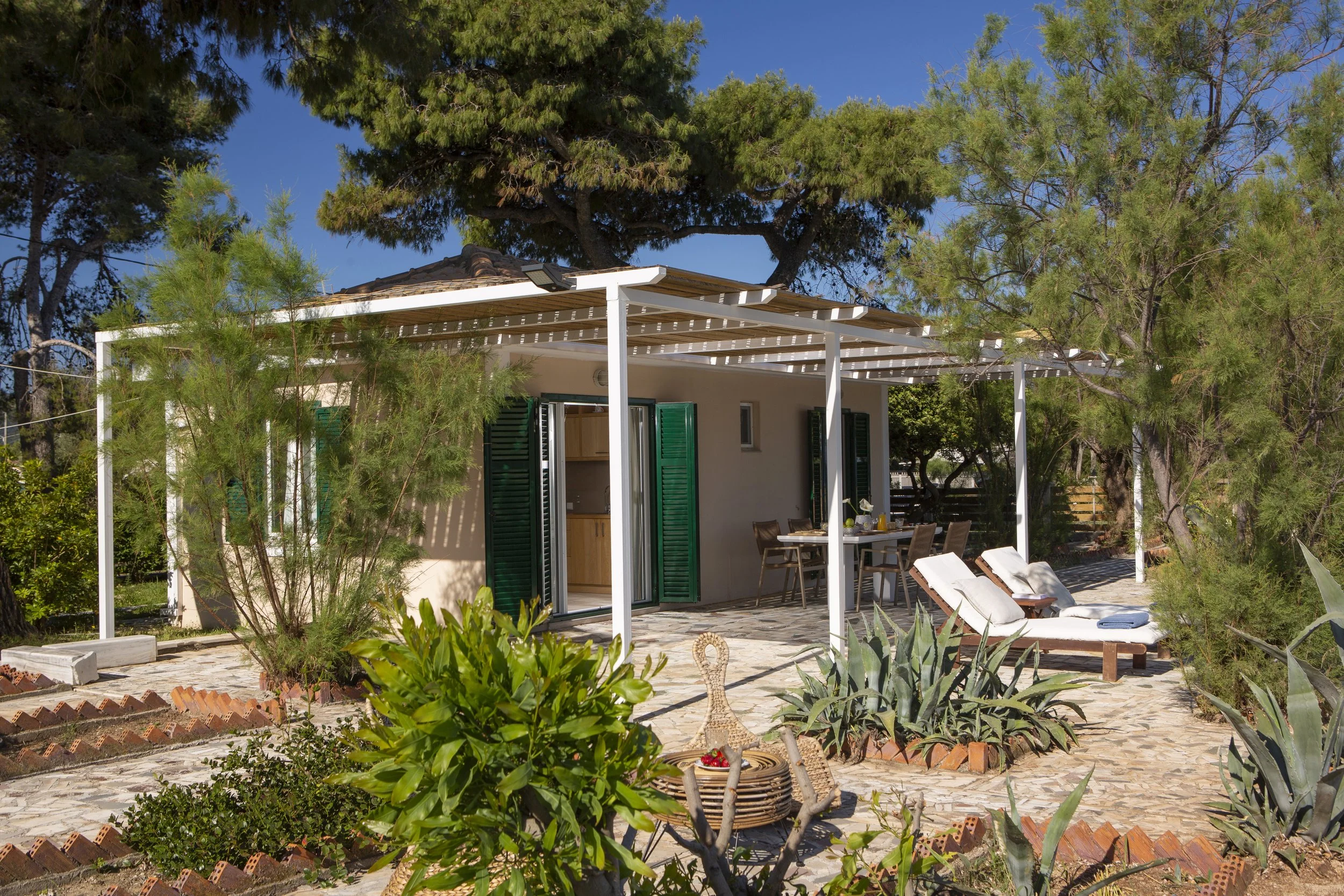 A backyard patio with a white pergola, surrounded by green plants and trees, with a small building featuring green shutters and an outdoor dining area with chairs and a table.