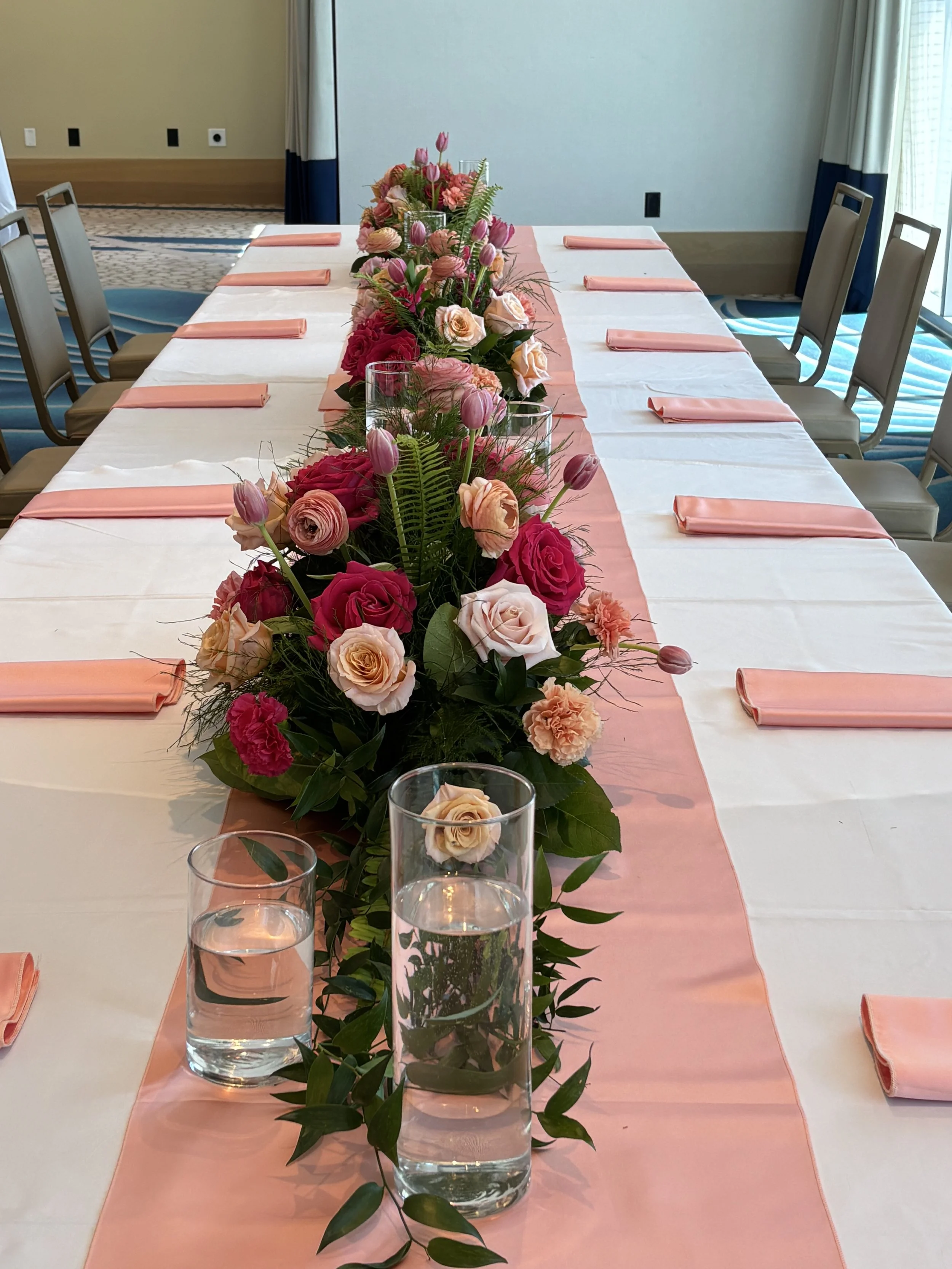 A long rectangular table decorated with a floral centerpiece featuring pink, peach, and red roses, greenery, and tulips. The table has a white tablecloth with a peach-colored runner and is set with pink napkins, glasses of water, and beige chairs aro