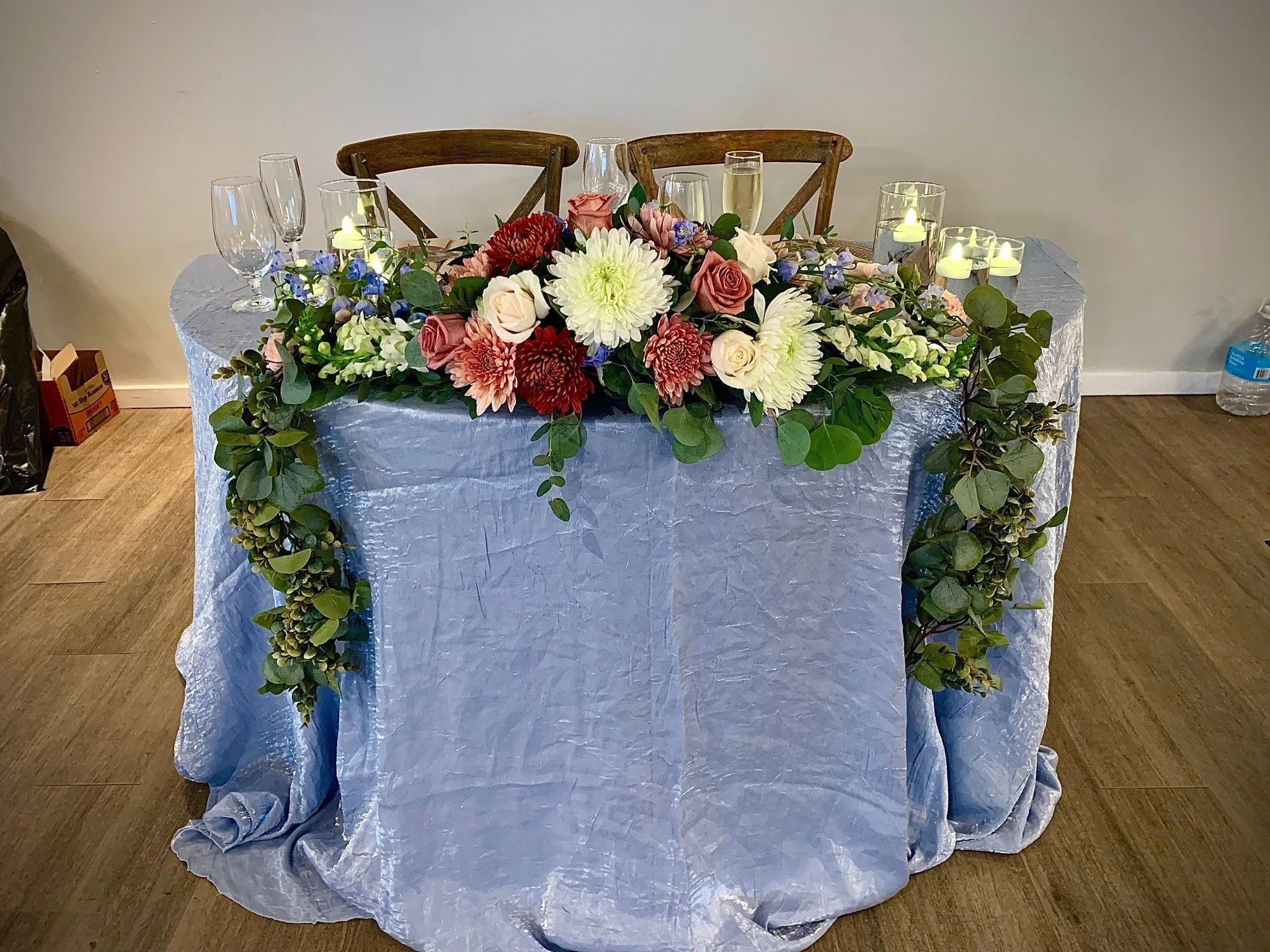 A banquet table decorated with a large floral arrangement of white, pink, and red flowers with green foliage, and surrounded by lit candles, set for a celebration or event.