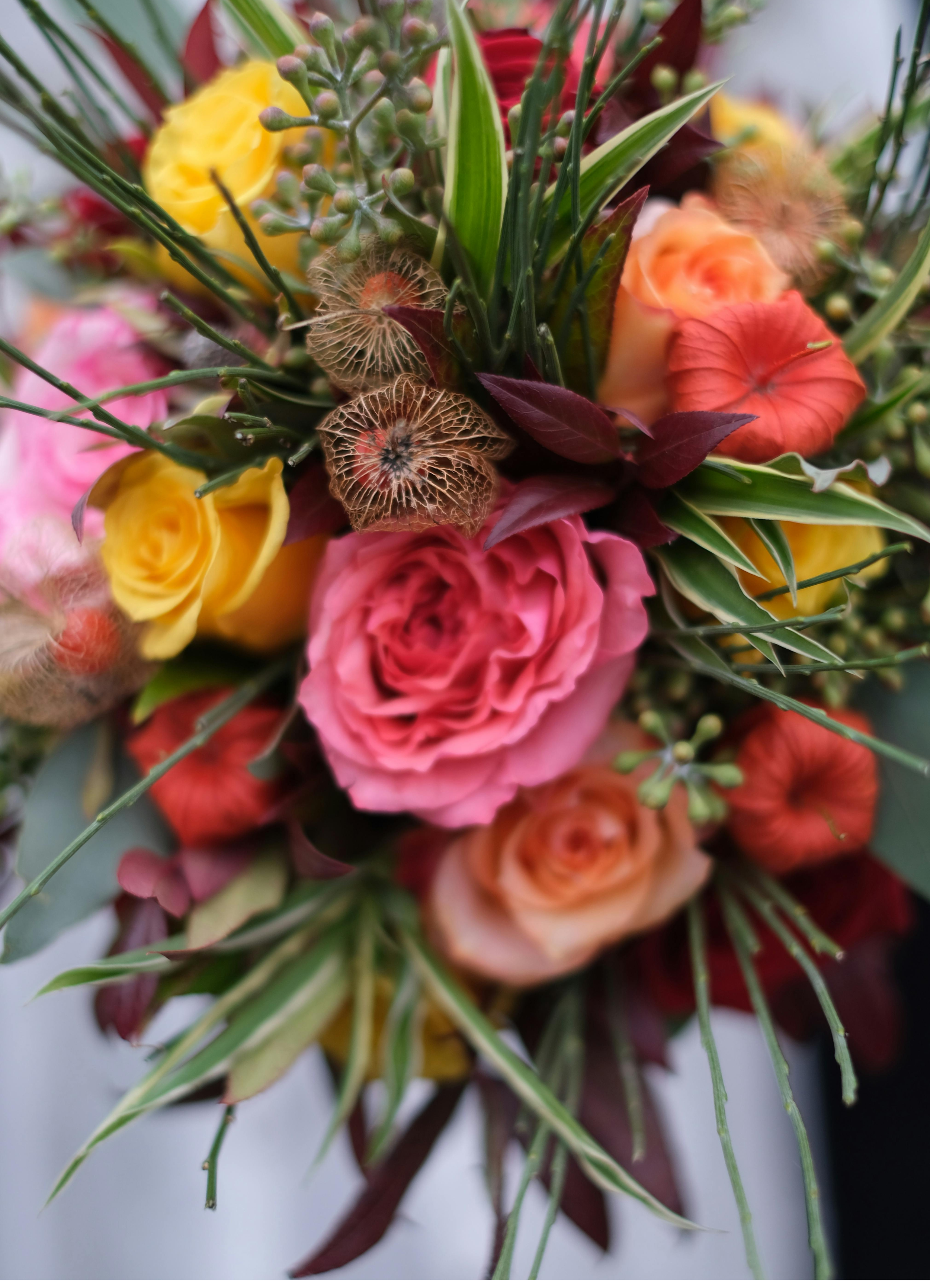 Close-up of a colorful flower bouquet including yellow, pink, red, and orange roses, green and variegated leaves, and delicate beige seed pods