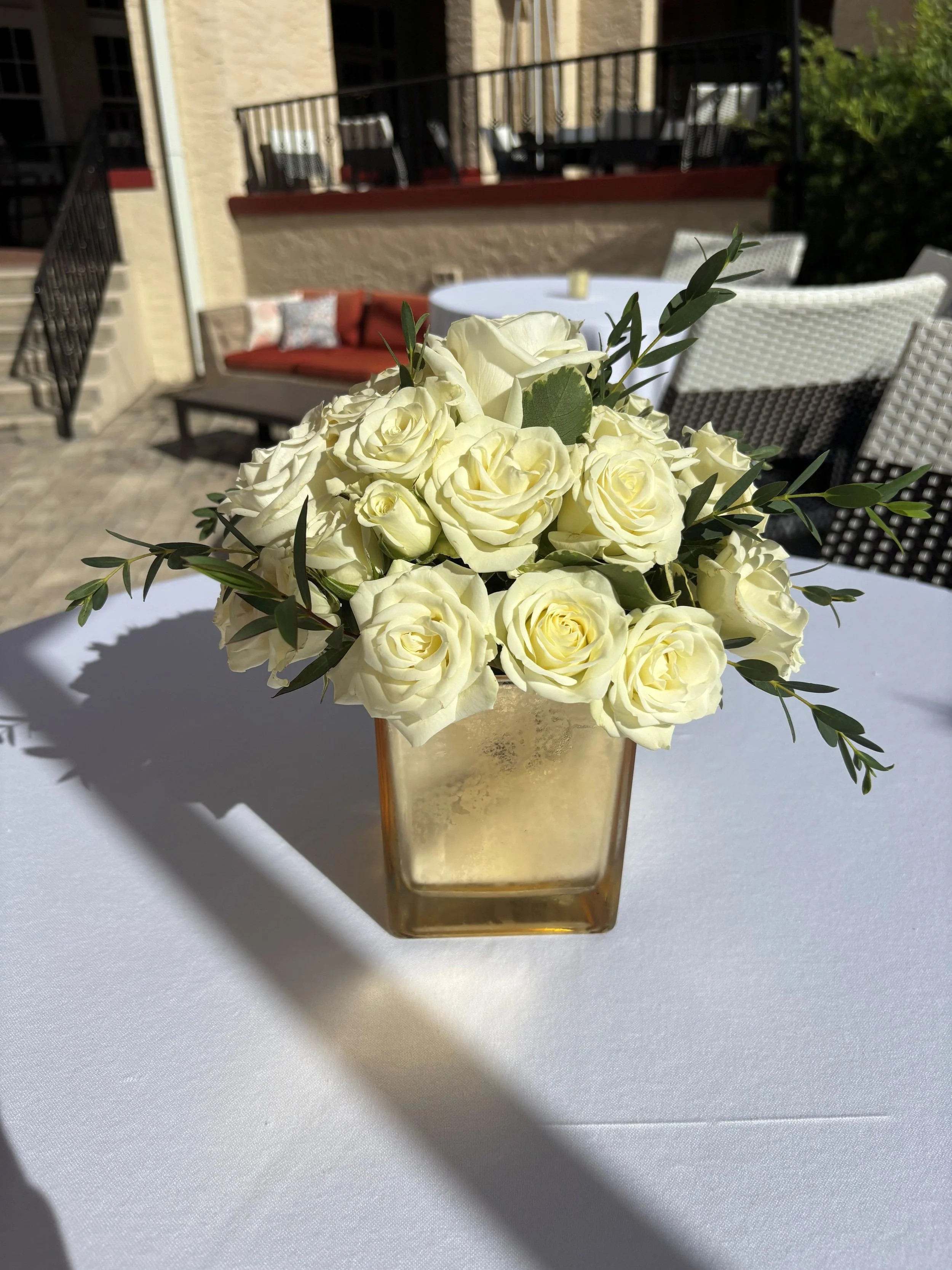 A bouquet of white roses and greenery in a golden vase on a white tablecloth