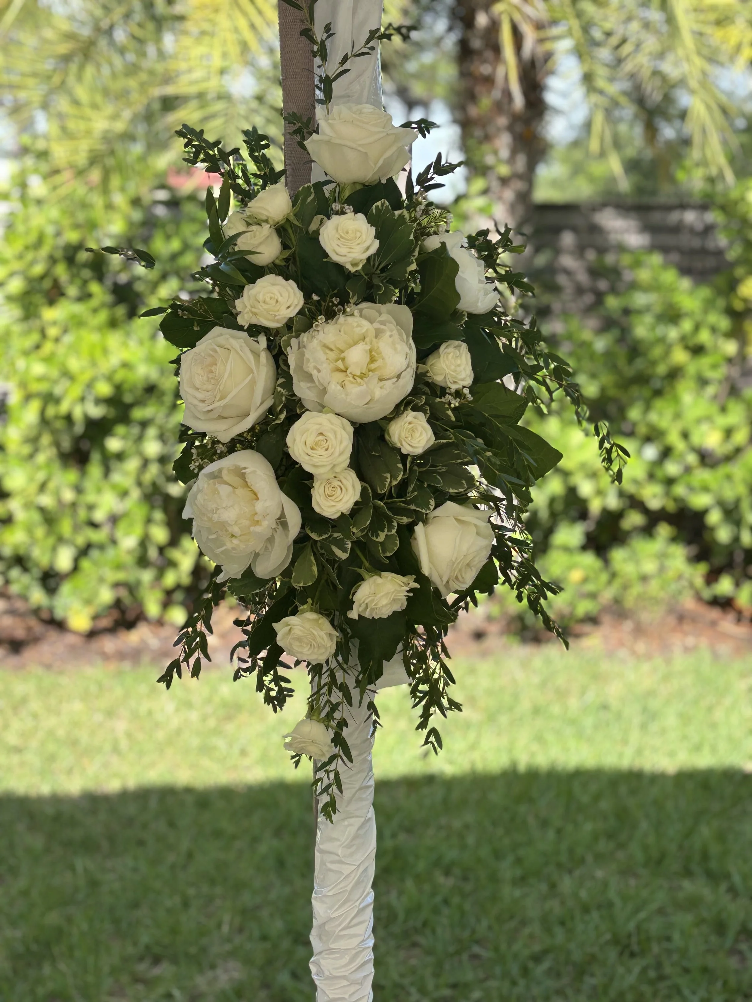 A bouquet of white roses and greenery hanging from a white cloth, against a background of trees and grass.