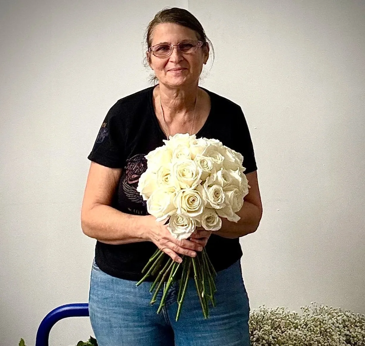 An older woman with glasses holding a bouquet of white roses, standing against a plain wall.