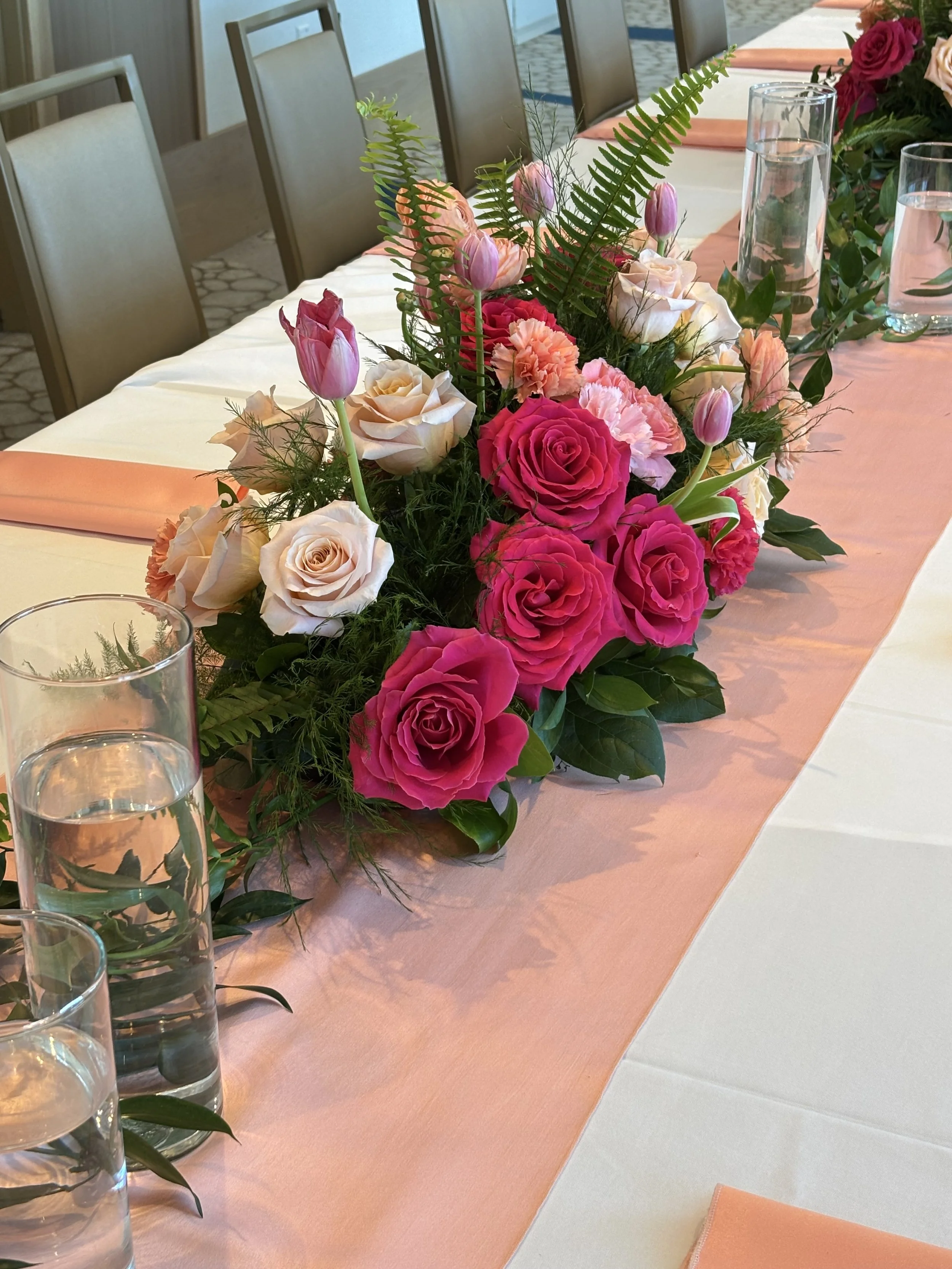Table decorated with a pink table runner and a floral centerpiece featuring pink, peach, and white roses, pink tulips, and greenery, with water glasses and orange napkins placed on the table.