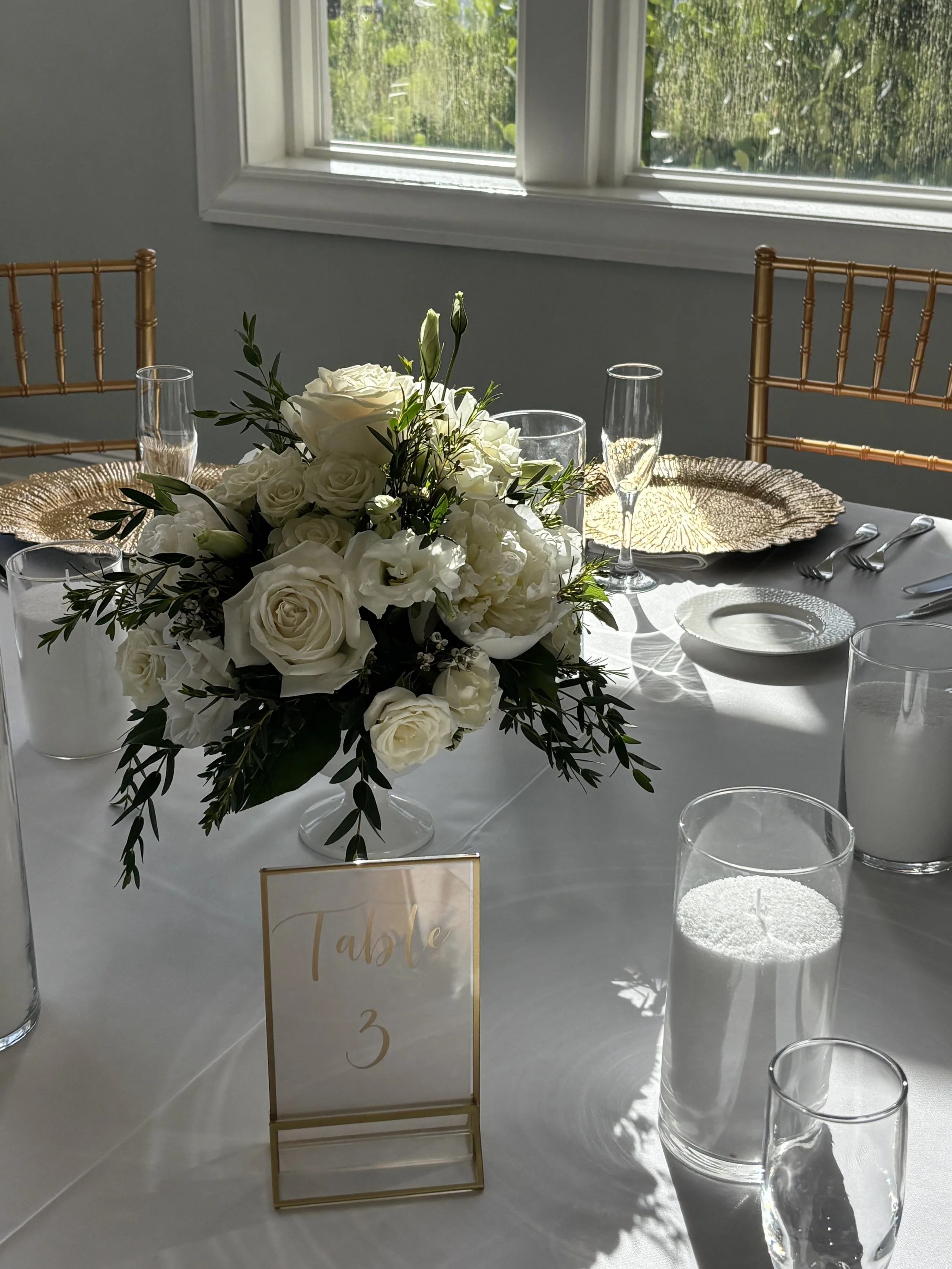 Elegant table setting with a bouquet of white roses and green foliage, gold chargers, glassware, and a table number sign reading 'table 3' in a bright room with a window showing greenery outside.