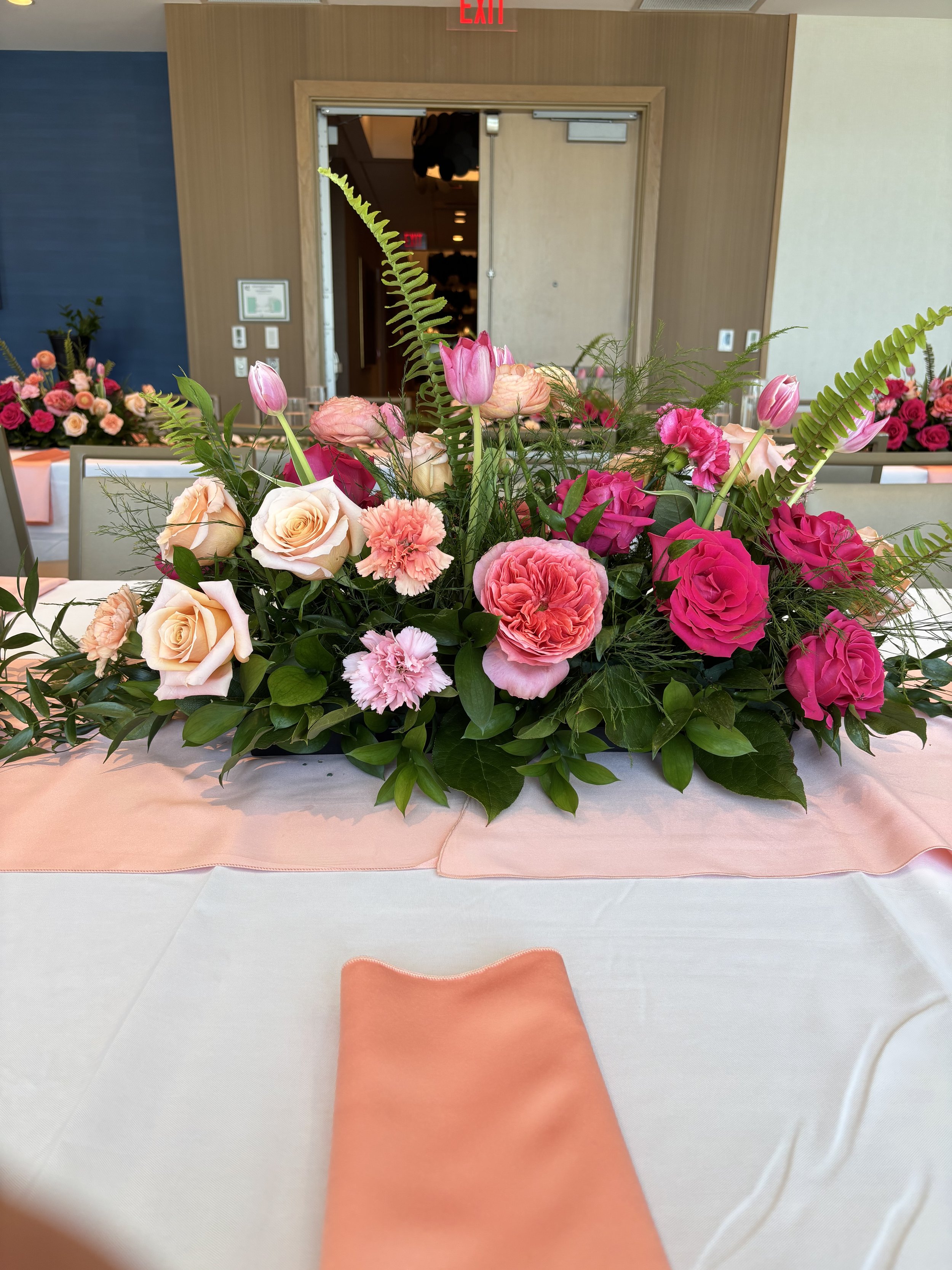 A floral centerpiece with pink roses, pink carnations, pink tulips, and greenery on a table decorated with a pink table runner, in a banquet hall with a wooden wall and an exit door in the background.