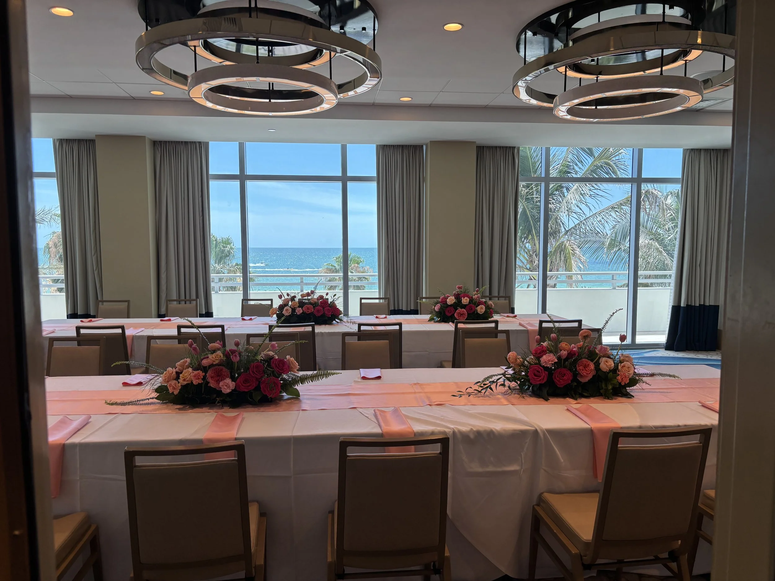 A room decorated with round modern chandeliers, large windows showing a view of the ocean, palm trees, white curtains, and a table with pink flowers and pink napkins for a celebration or event.