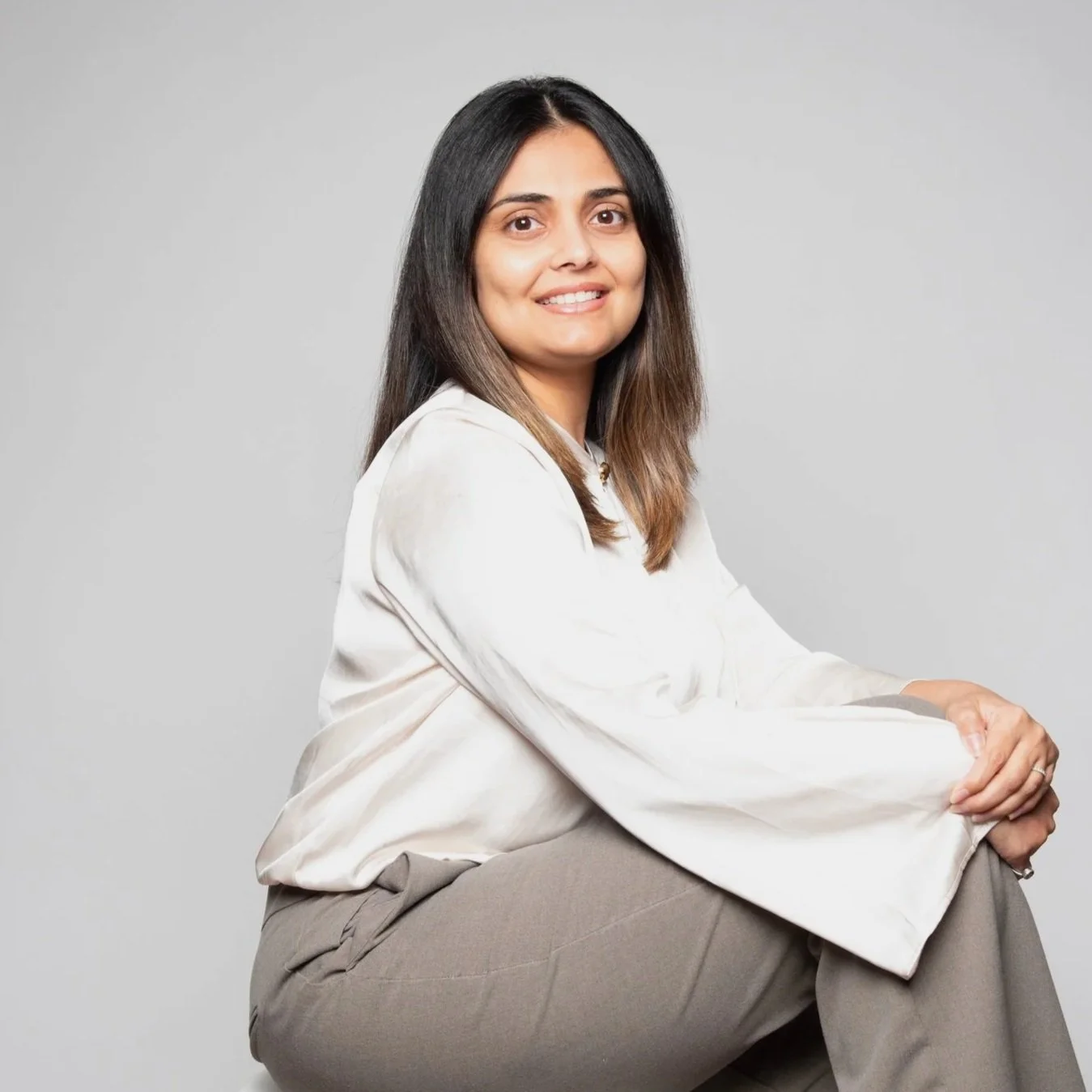 A woman with shoulder-length brown hair, wearing a white satin blouse and gray pants, sitting on a white stool against a plain light gray background, smiling at the camera.