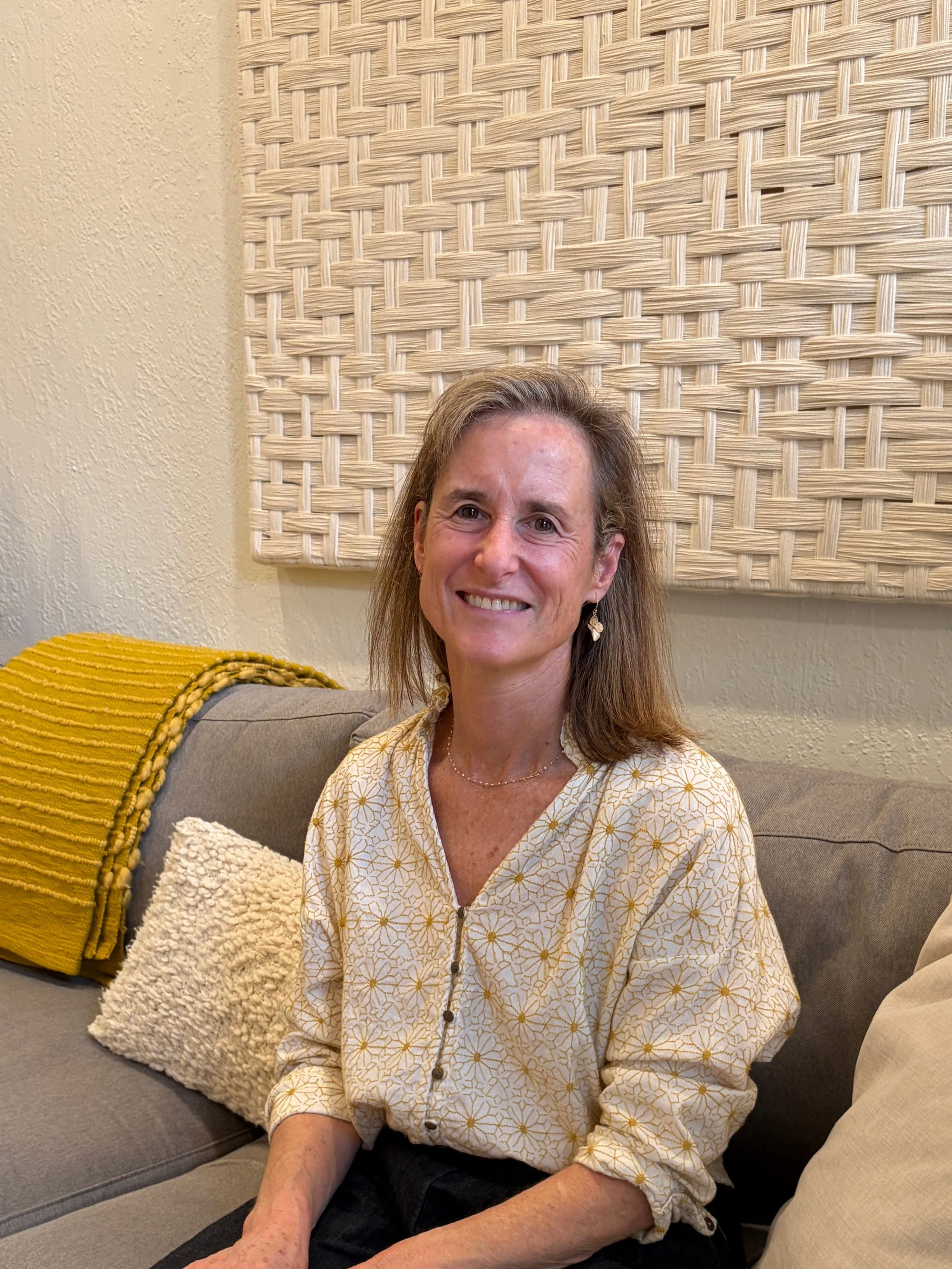 A woman sitting on a gray couch, smiling, wearing a cream-colored blouse with yellow floral patterns, with a yellow pillow and cream textured blanket on her left, and a woven artwork on the wall behind her.