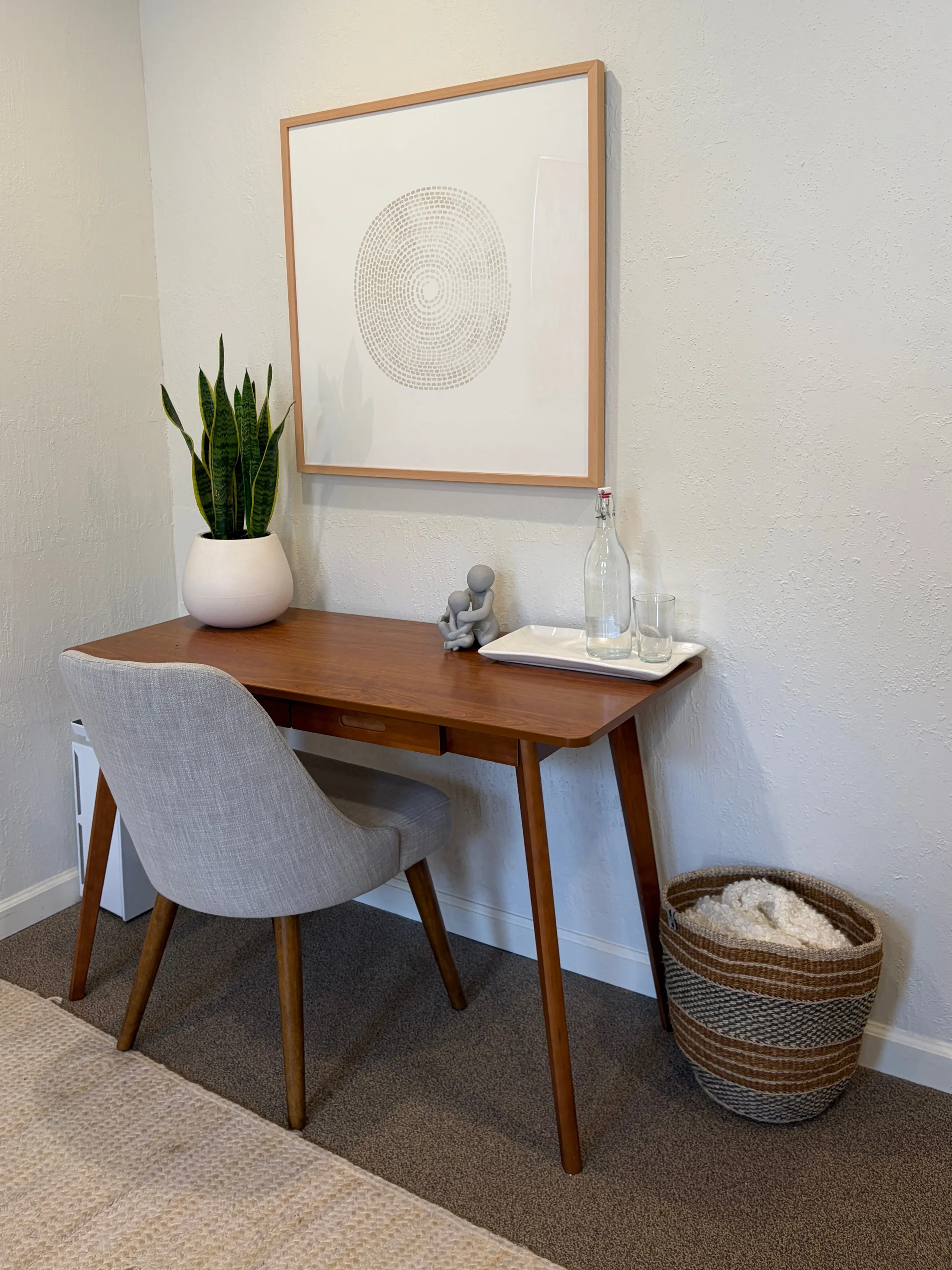 A wooden desk with a gray upholstered chair, a tall potted snake plant, a decorative sculpture, and a tray with a glass bottle, glass, and a small container. There is a framed modern art piece and a woven basket on the floor.