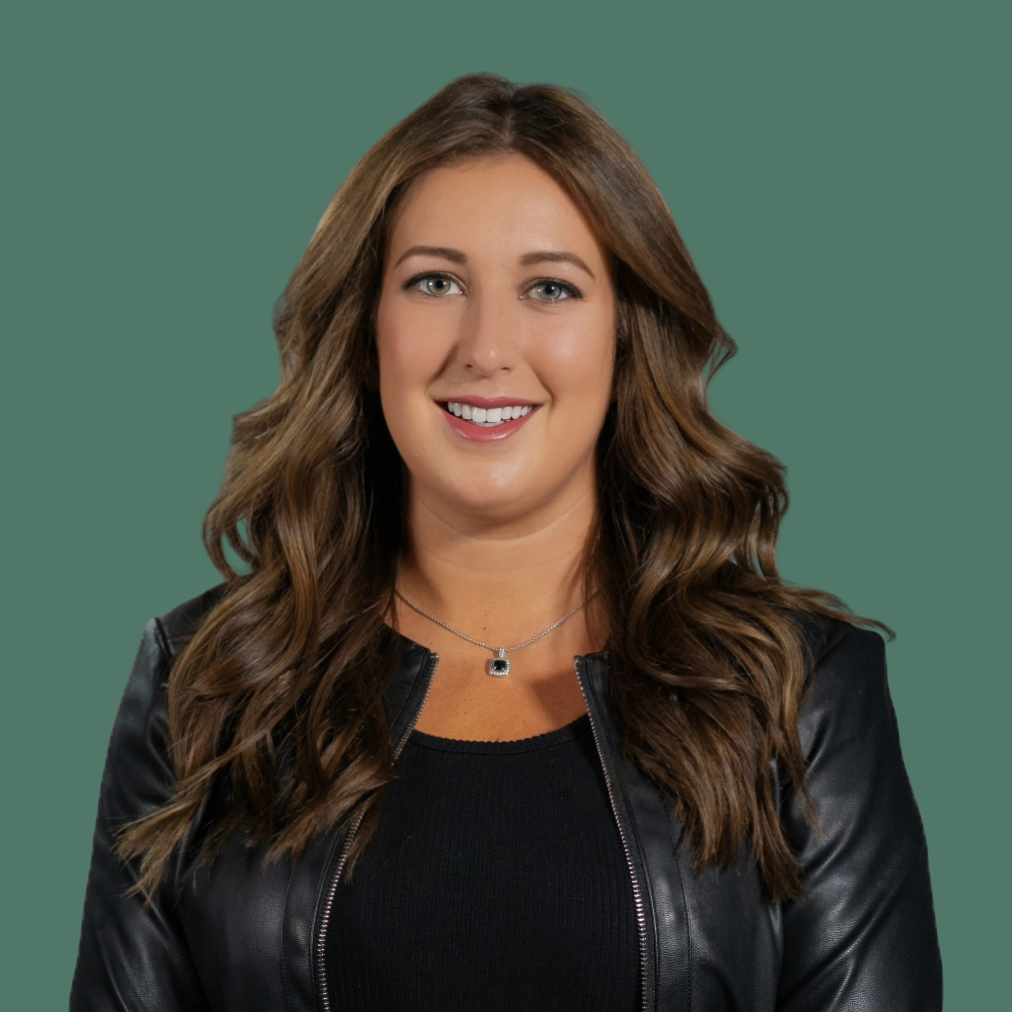Portrait of a smiling woman with long wavy brown hair, wearing a black top, a black leather jacket, and a silver necklace with a square pendant, against a plain background.