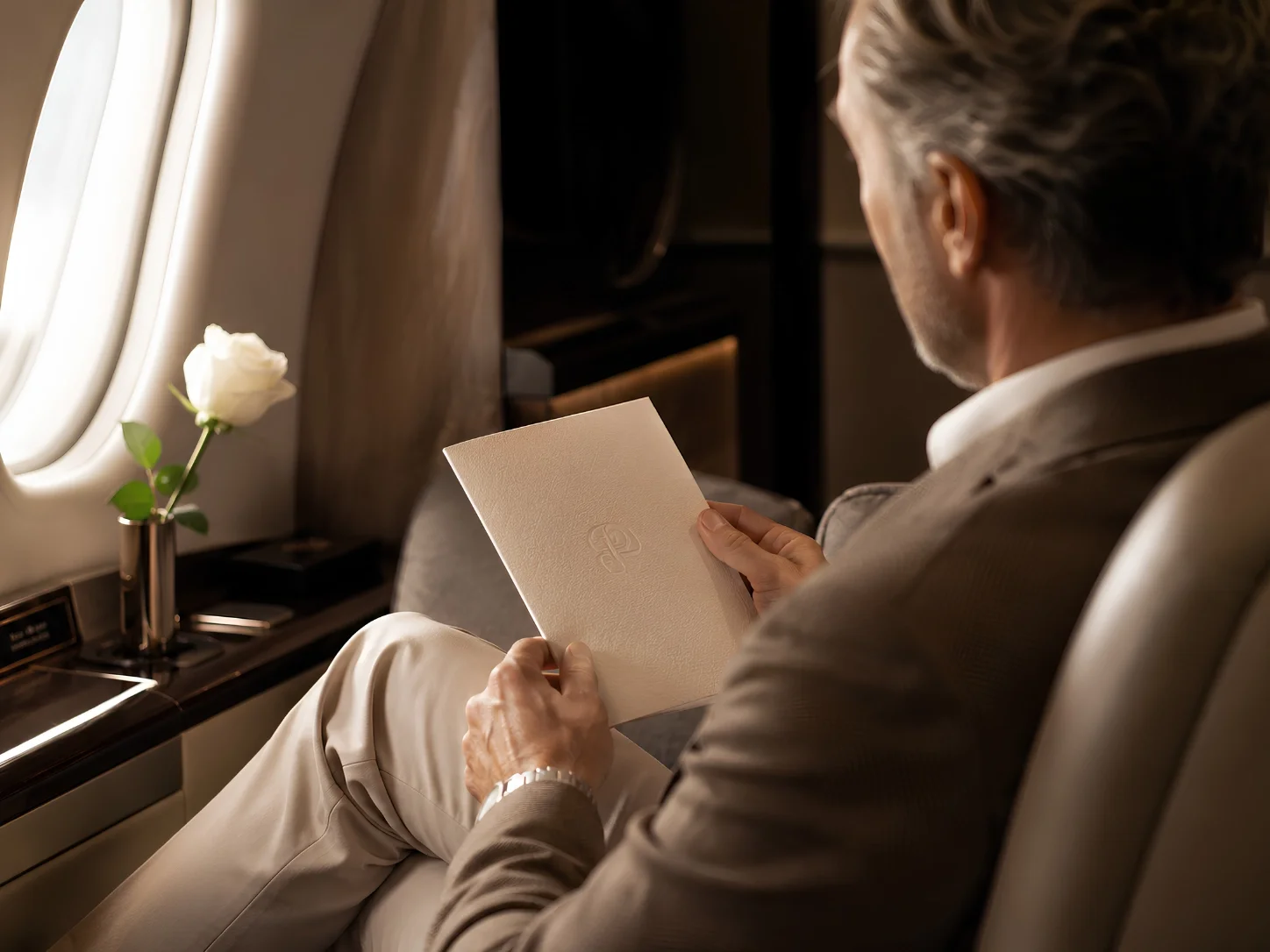 En man som sitter i ett flygplansäte och läser en vit folder, med en vit ros i en vas på fönsterbordet intill