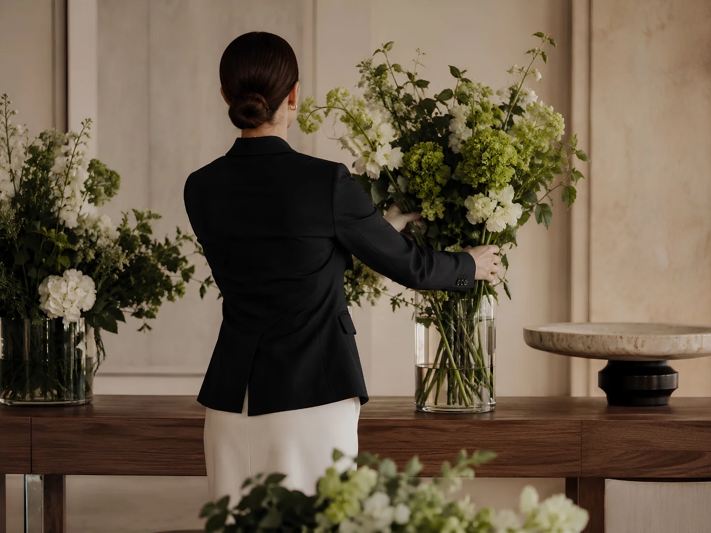 En kvinna i svart kavaj som arrangerar vita och gröna blommor i ett glasvasar på ett bord.
