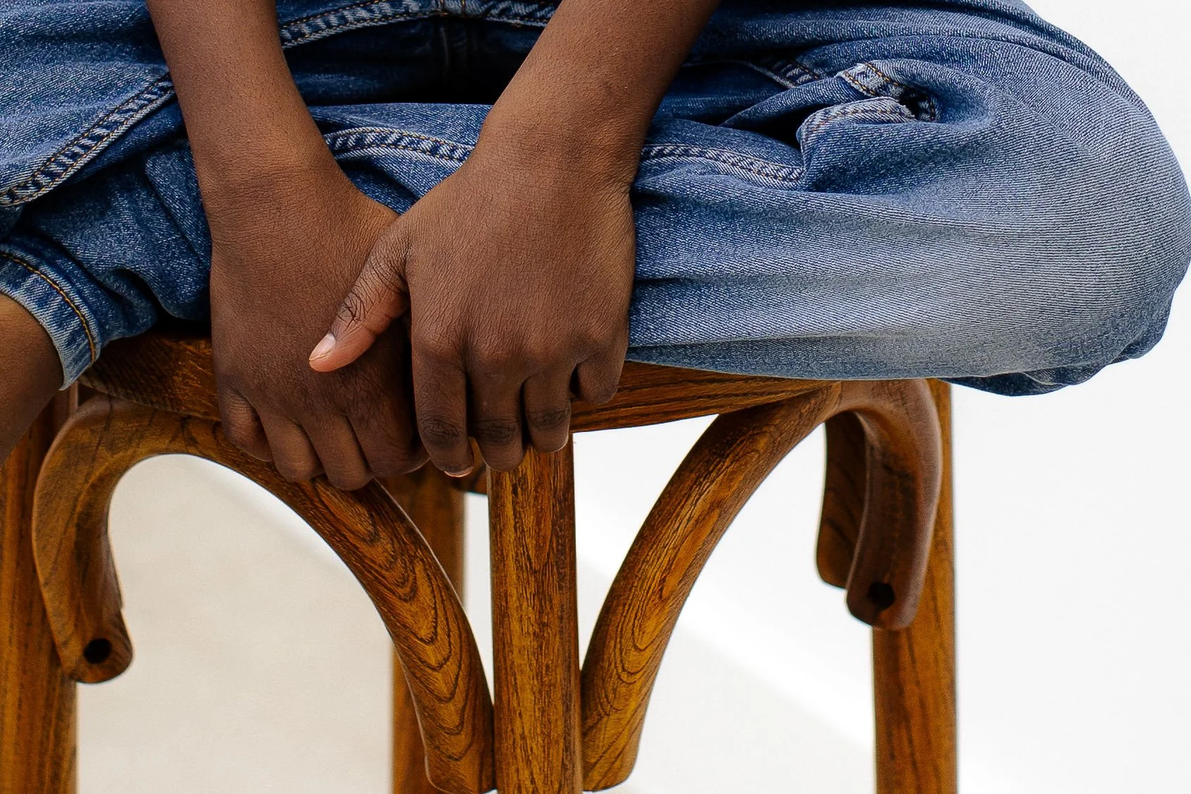 A person with dark skin wearing blue jeans, sitting cross-legged on a wooden chair, with their hands clasped together resting on their lap.