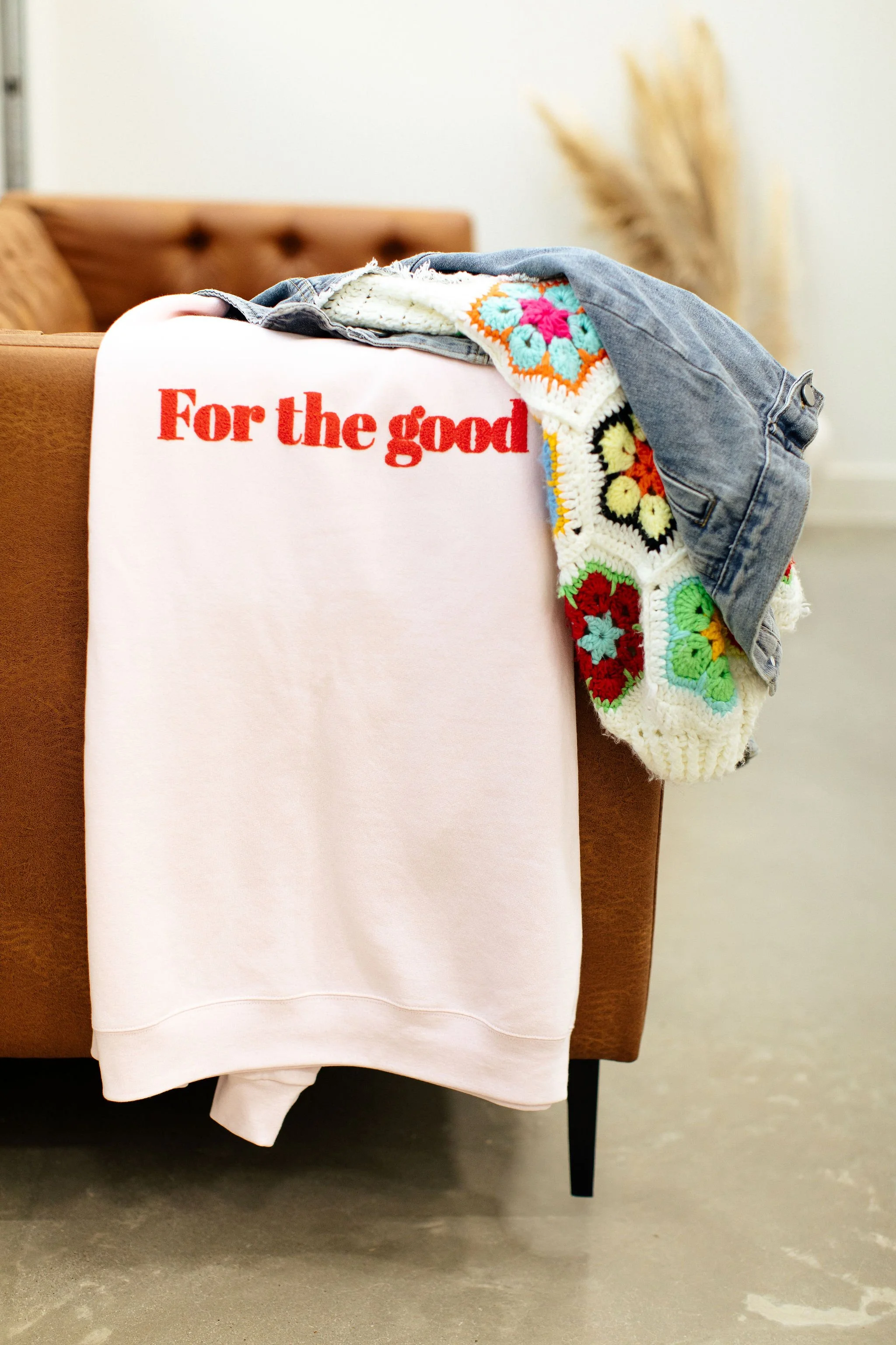 Clothes draped over a piece of furniture, including a pink sweatshirt with red text, a multicolored crochet blanket, and denim jeans, in a cozy indoor setting.