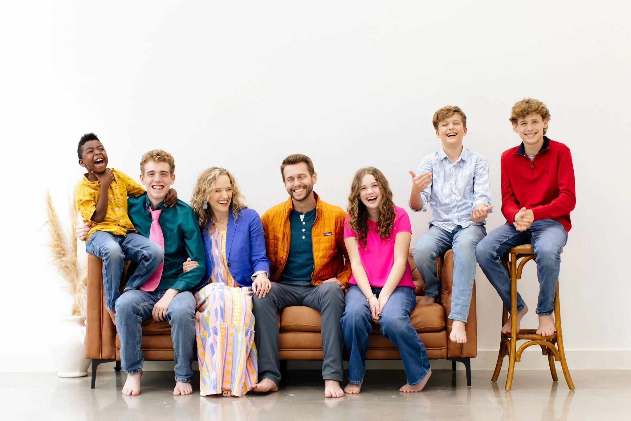 A diverse group of seven young people sitting and standing on and around a brown sofa, laughing and smiling indoors with a plain white background.