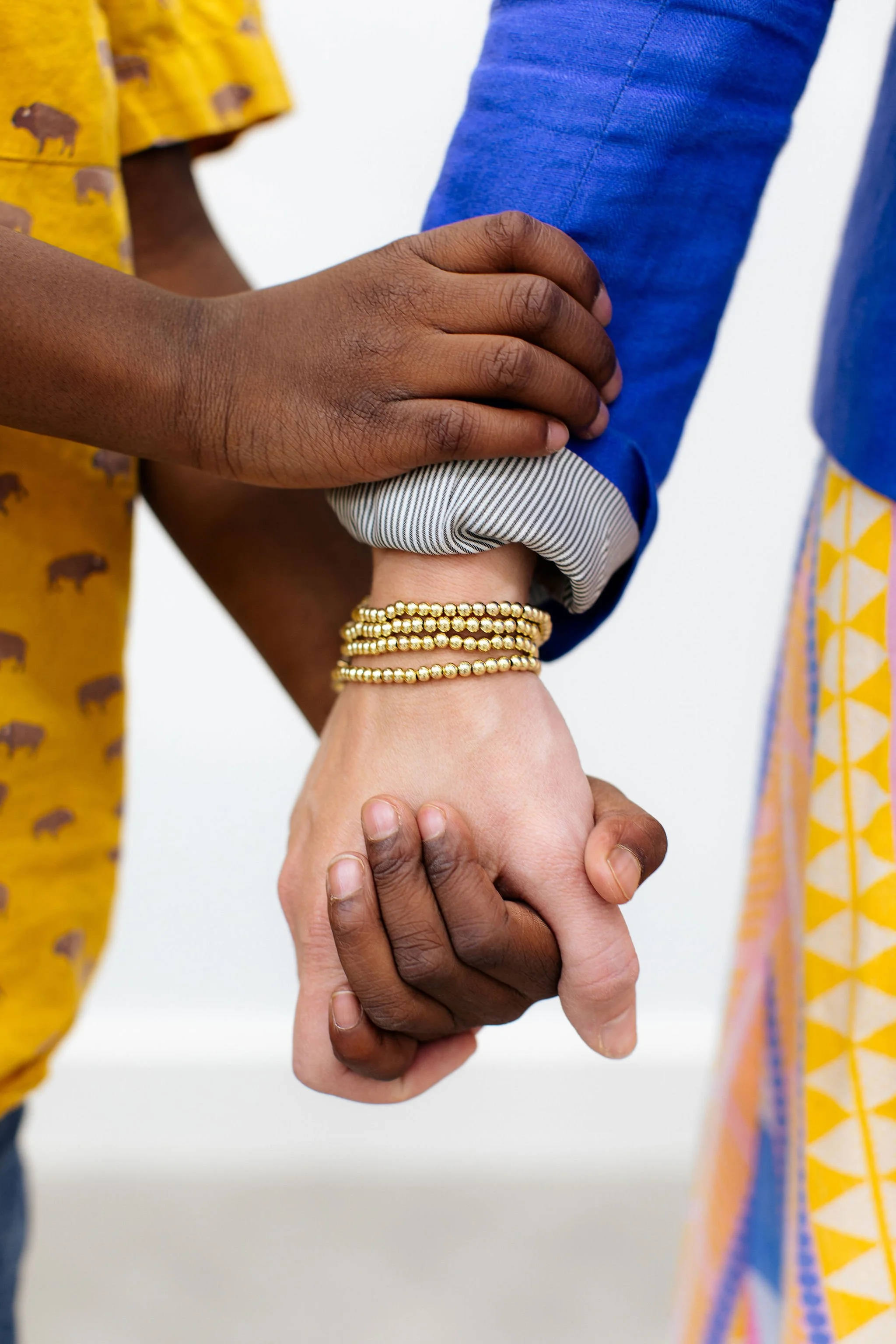 A close-up of two people holding hands, one with darker skin wearing gold bracelets and the other with lighter skin. The person with darker skin is gently resting their hand on the other's arm, which is dressed in a blue cuffed shirt. The background is plain and white.
