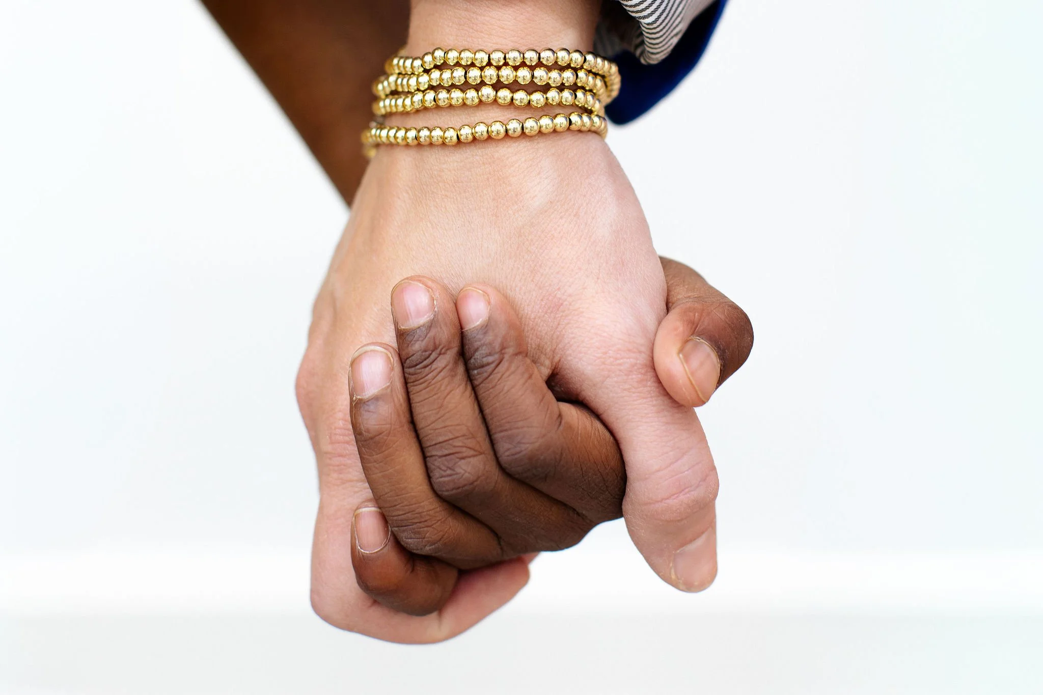 Close-up of two people holding hands, one with darker skin and wearing gold bead bracelets, and one with lighter skin.
