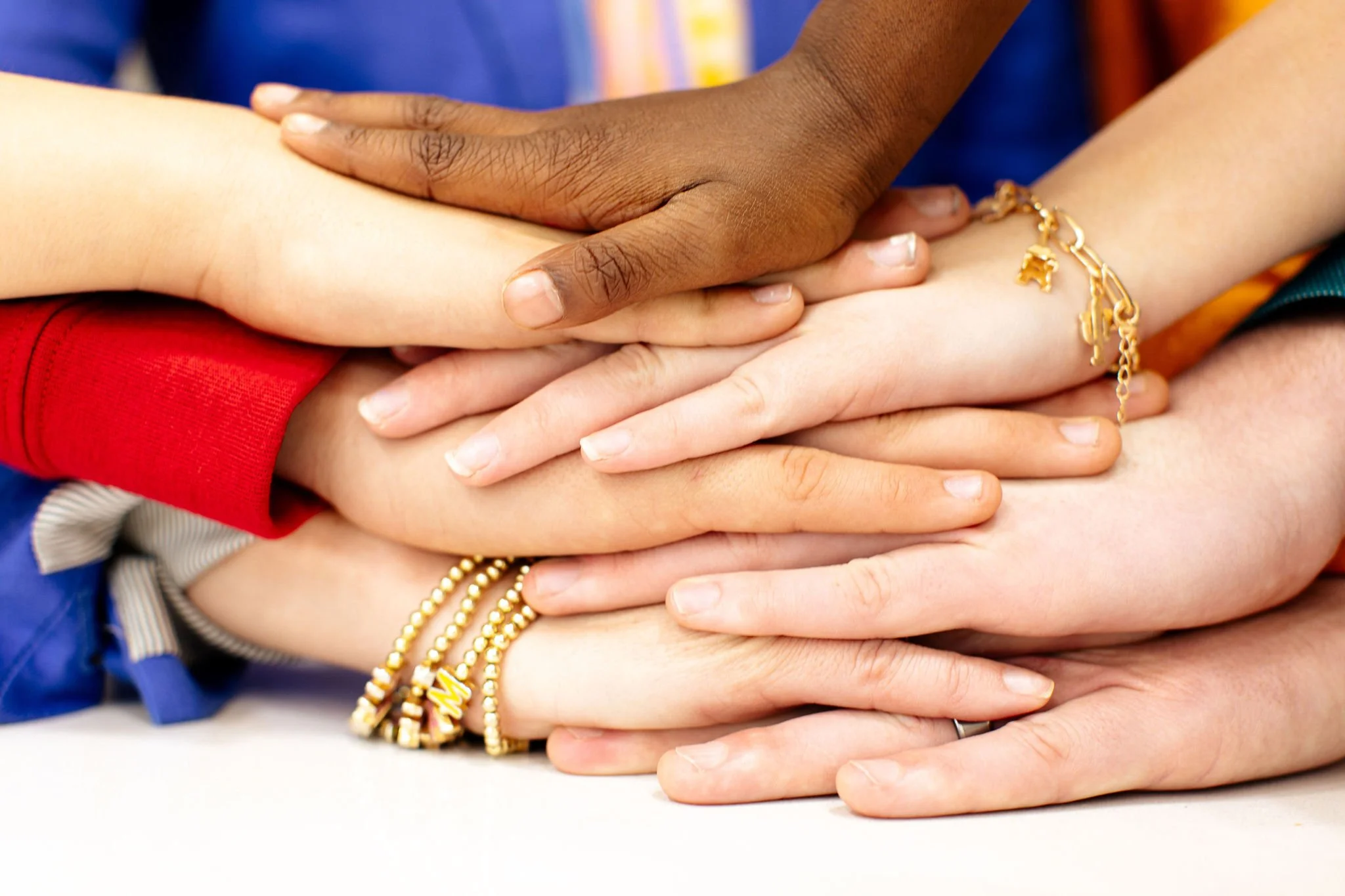 A group of diverse hands stacked together, some wearing gold bracelets and rings.
