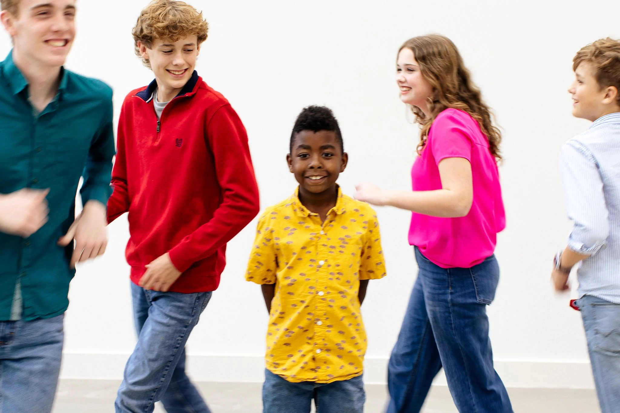 A group of five children smiling and interacting with each other in a room with a plain white wall. The children are dressed casually in colorful clothes.