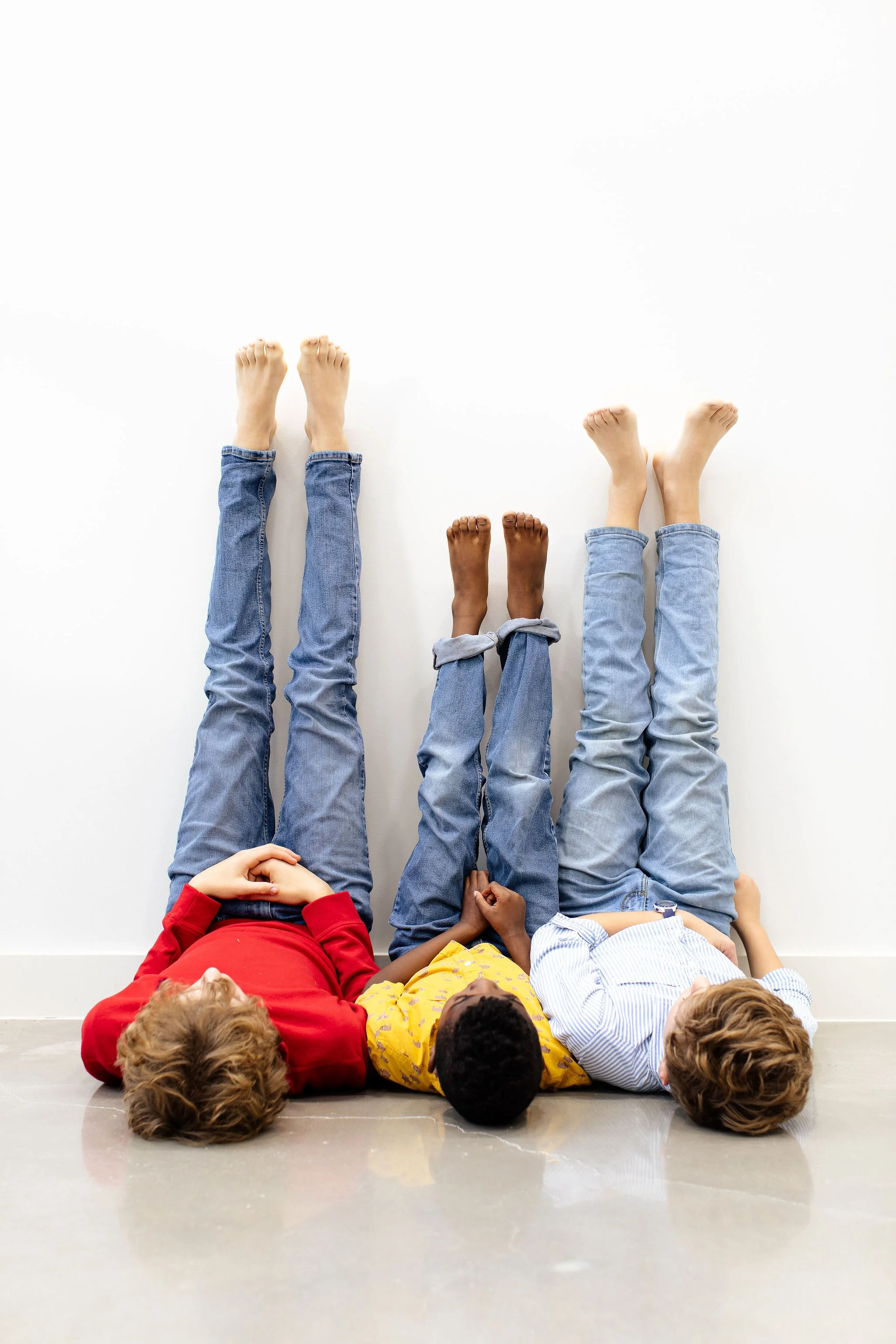 Three people lying on the floor with their heads against a white wall and their legs raised vertically, wearing jeans and different colored tops.