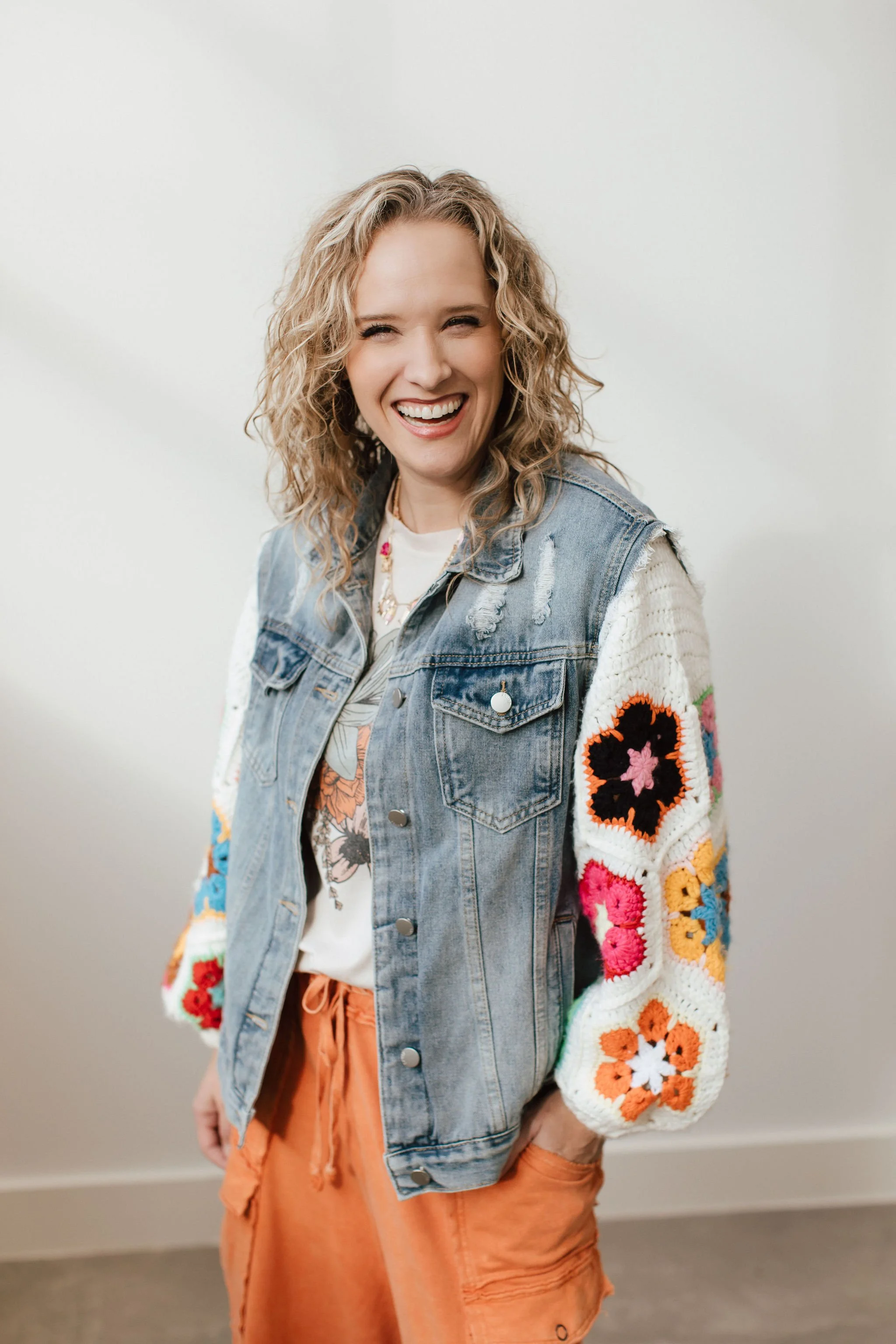 A woman with curly blonde hair smiling and wearing a denim vest with floral and crochet sleeves, a white t-shirt, and orange pants, standing against a plain white wall.