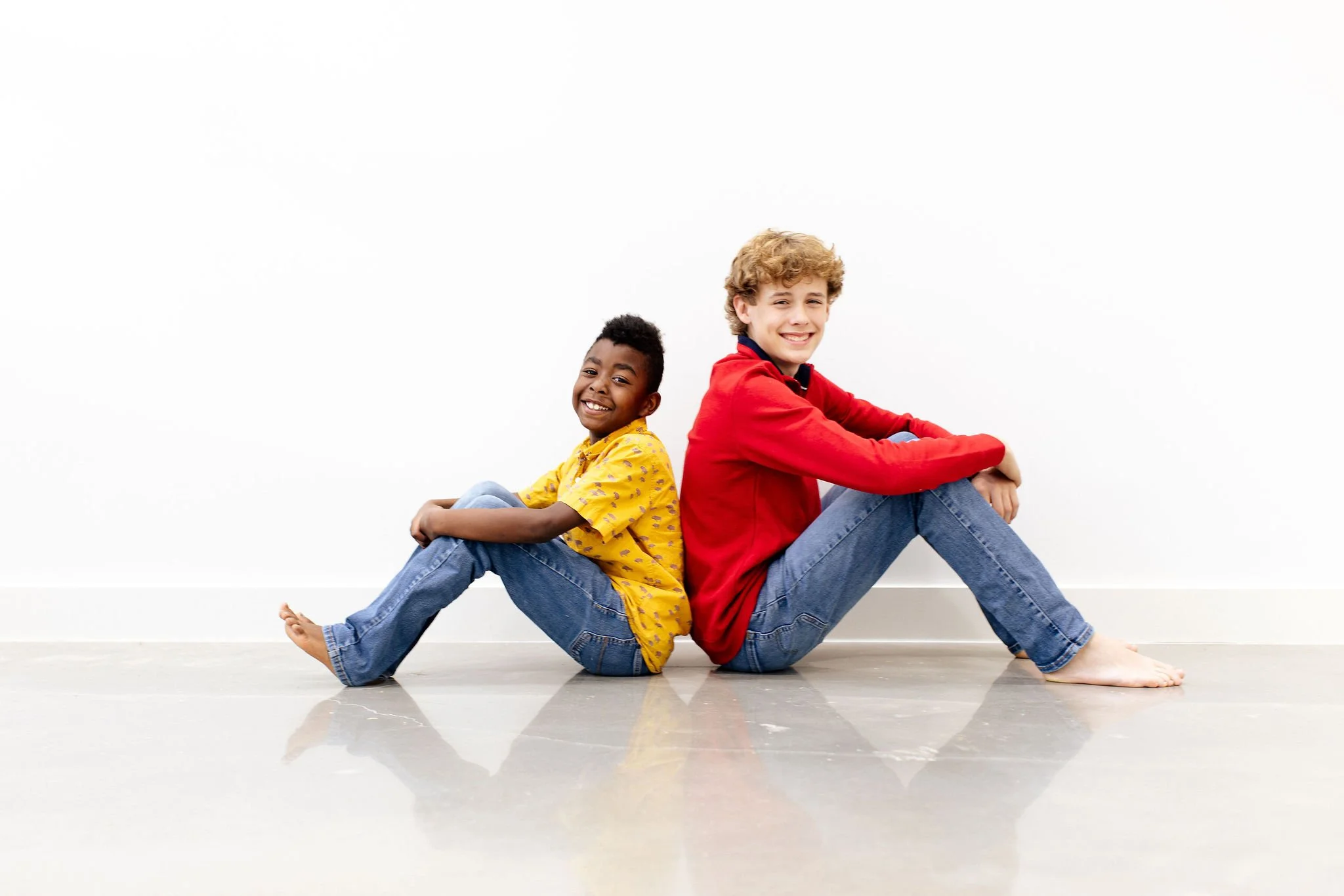 Two boys sitting on the floor against a white wall, smiling. One boy wears a yellow shirt and jeans, the other wears a red shirt and jeans.
