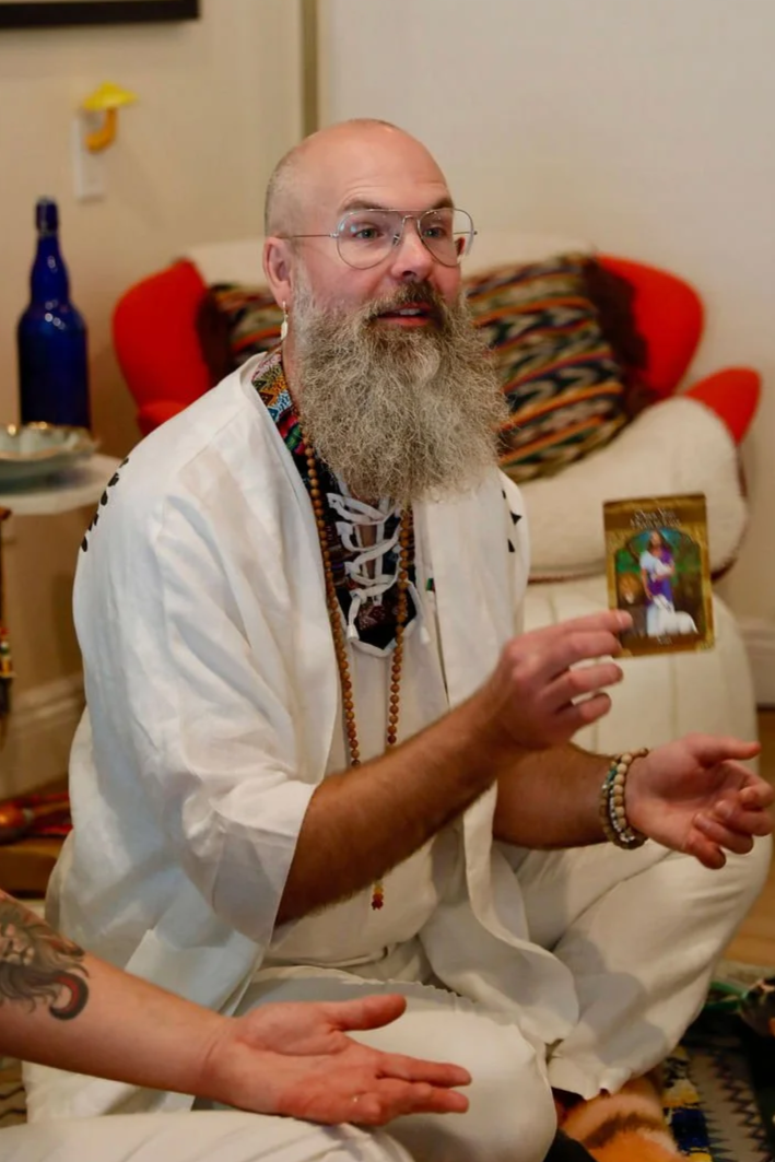 A man with glasses, a long beard, and a bald head sits on the floor indoors, holding a tarot card. He wears a white shirt, beaded necklaces, and bracelets, surrounded by a colorful, bohemian setting.